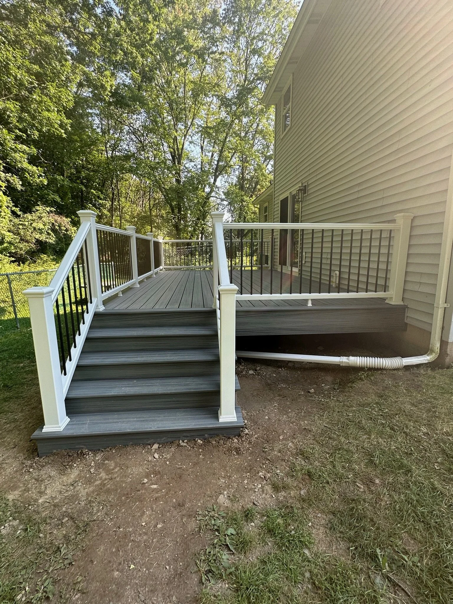 A newly built wooden deck with steps leading up to it, attached to a house with beige vinyl siding. The deck features white and black railings and is surrounded by trees and greenery.