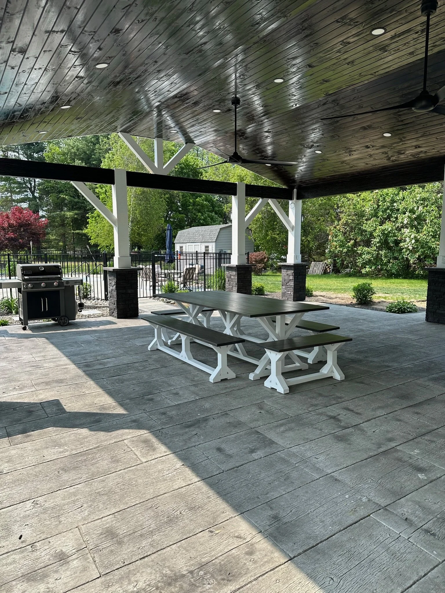 Covered outdoor patio with a wooden ceiling, ceiling fans, a picnic table, a barbecue grill, and surrounding greenery.