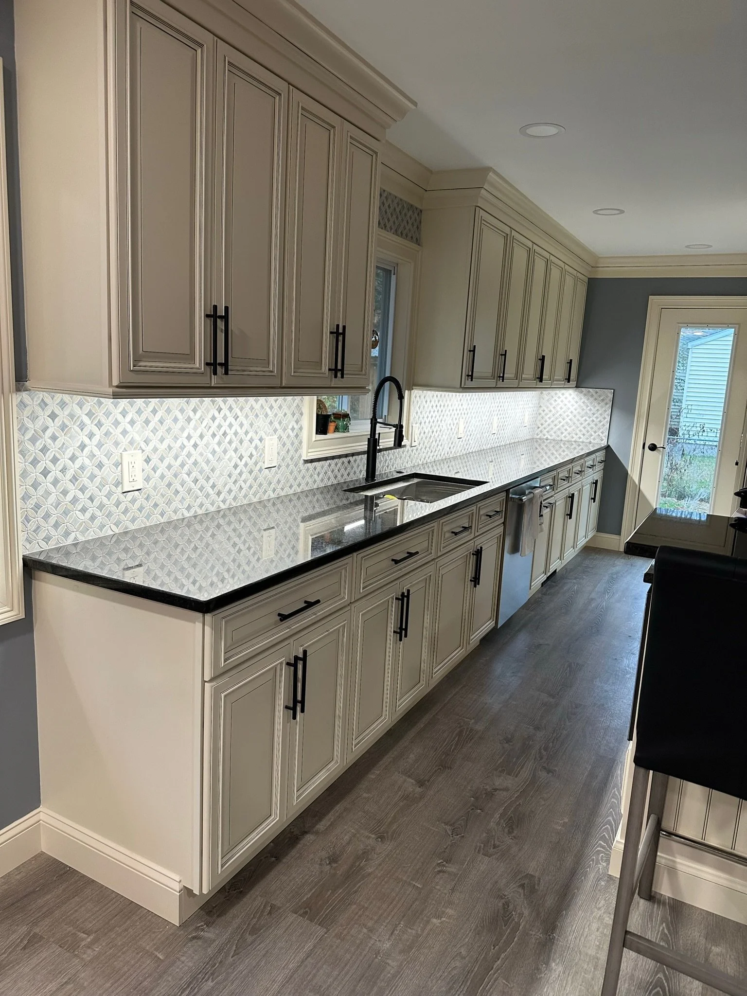 Modern kitchen with cream-colored cabinets, black hardware, granite countertop, white patterned backsplash, black faucet, and a door leading outside.