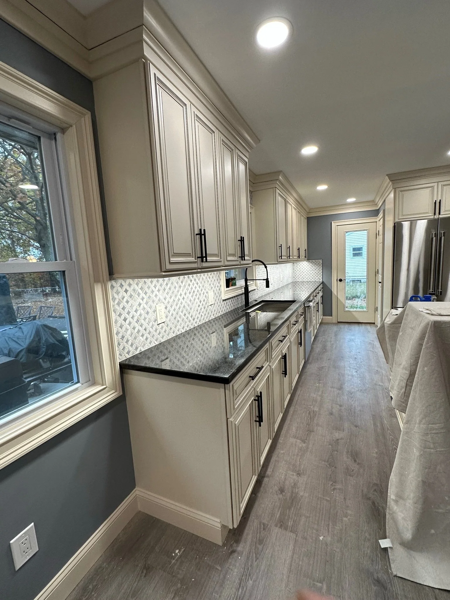 Modern kitchen with beige cabinets, black countertop, patterned backsplash, stainless steel refrigerator, and gray wooden flooring.