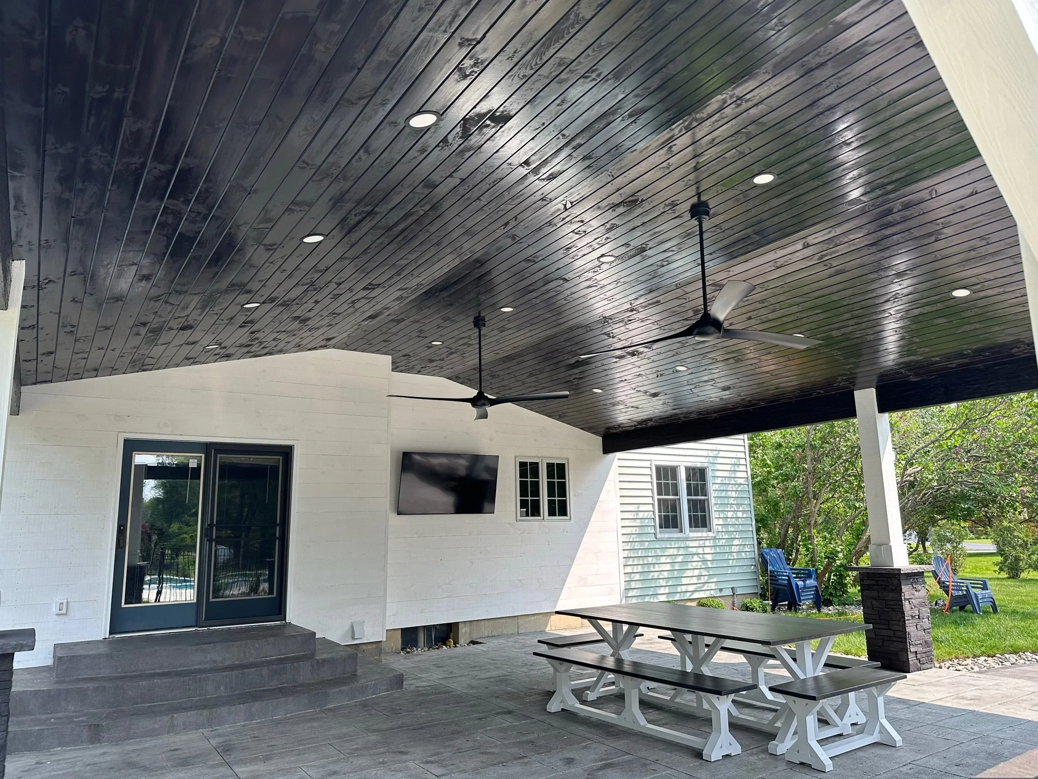 Covered outdoor patio area with a dark wood ceiling, ceiling fans, a white wall with a television, a glass door, window, a picnic table with benches, and some chairs outside near a grassy yard with trees.