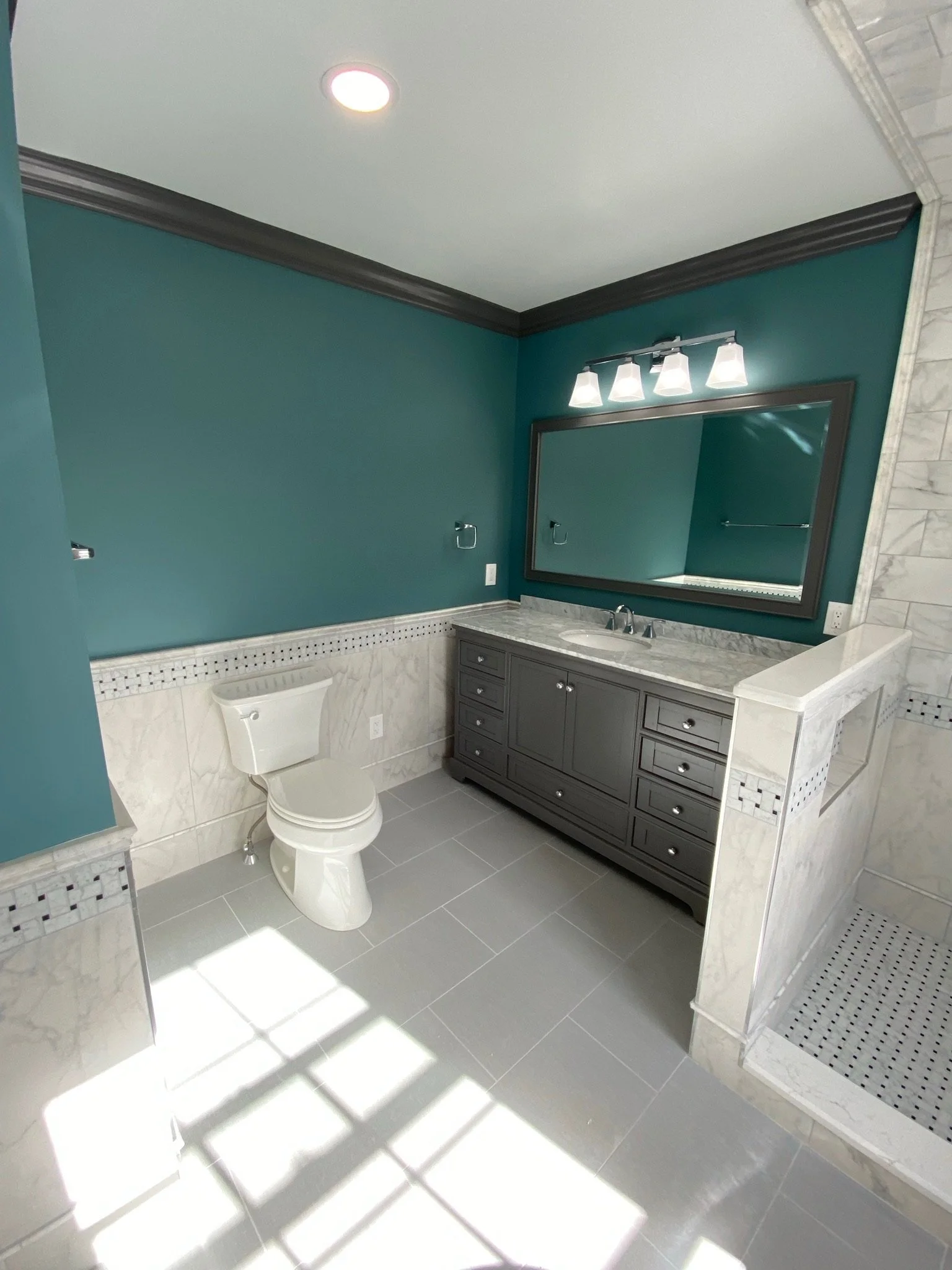 A modern bathroom with teal walls, a large mirror above a gray vanity with a marble countertop, a toilet, and a walk-in shower with marble tiles and a mosaic tile border. There is a light fixture above the mirror and sunlight coming through a window