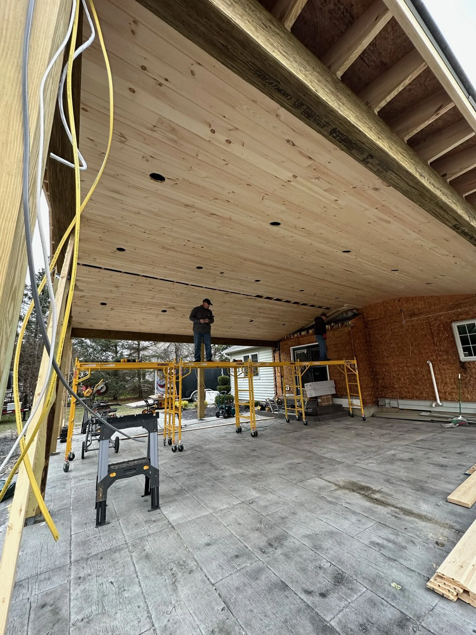 Construction workers installing a wooden ceiling under a house extension, with scaffolding and construction tools around.