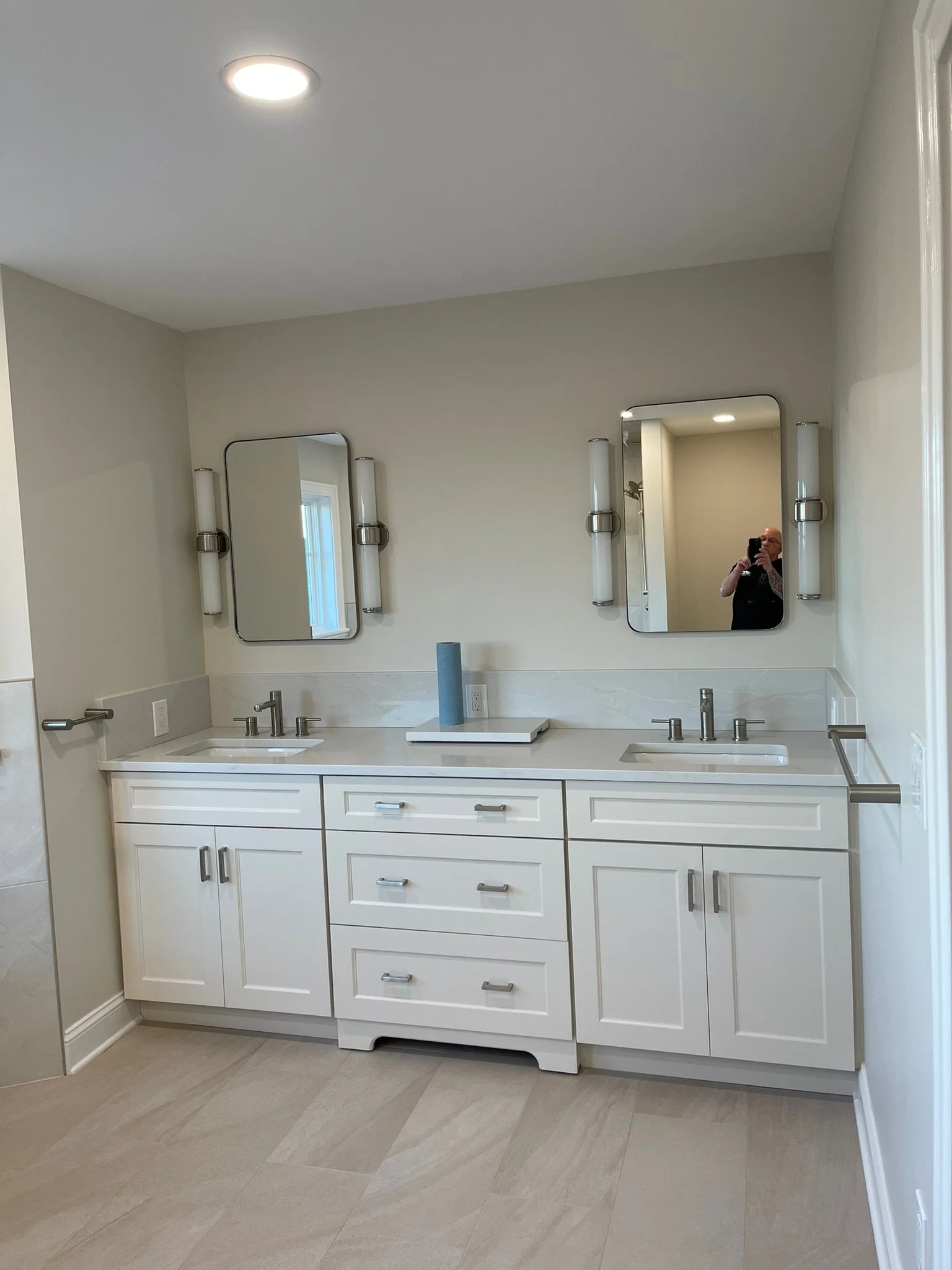 A modern bathroom vanity with a double sink, white cabinetry, and two mirrors with horizontal lights on either side. There is a small blue towel on a tray between the sinks, and the bathroom has light-colored tile flooring and neutral walls.