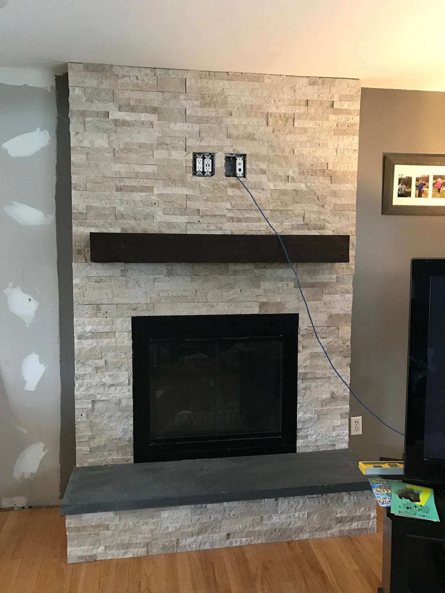 Living room with a stone fireplace, black mantel, and unarranged electrical outlets on the wall, with a wire hanging from one of the outlets.