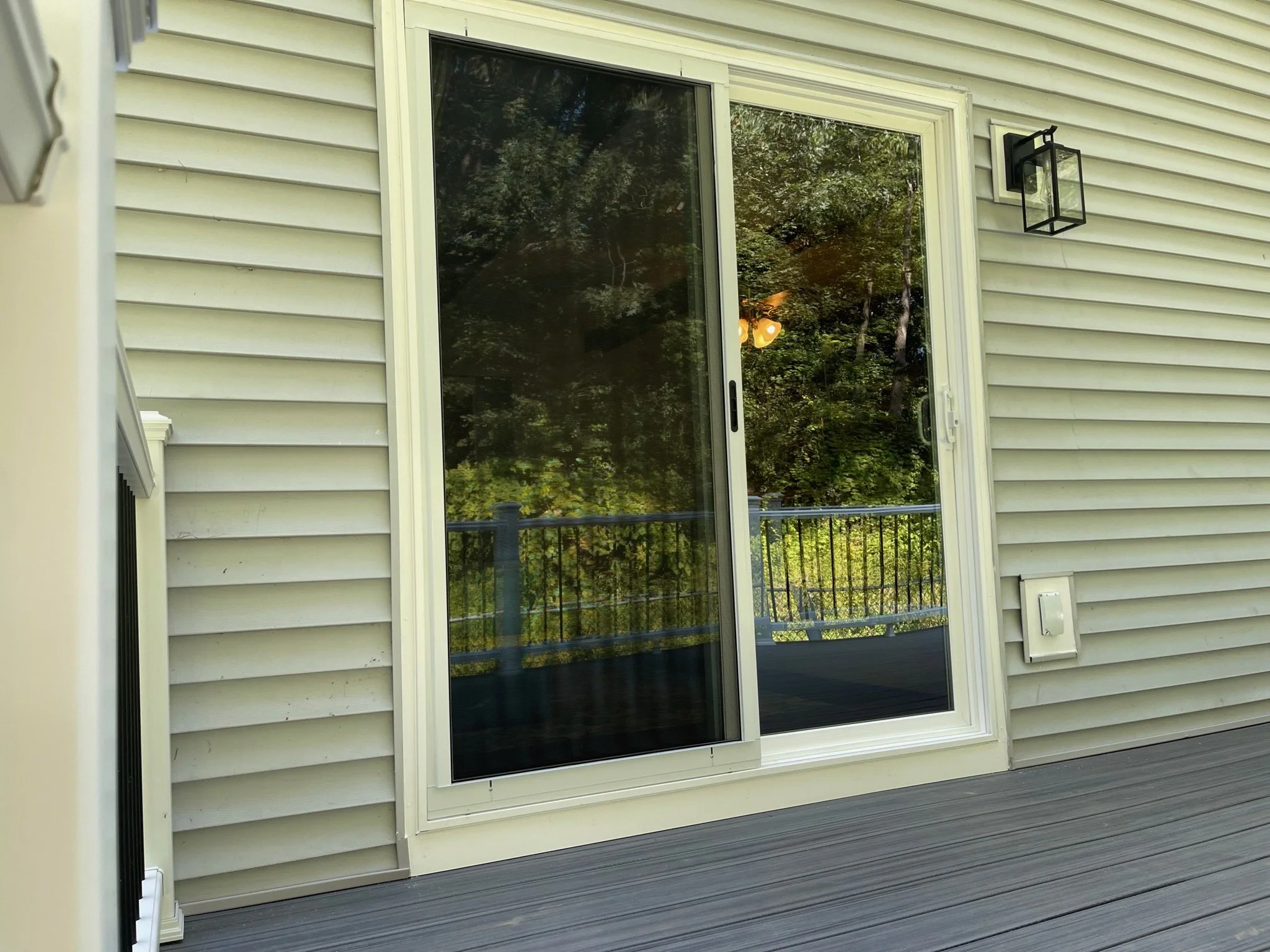 Exterior view of a house showing a sliding glass door, beige vinyl siding, an outdoor lantern, and a deck with a railing.