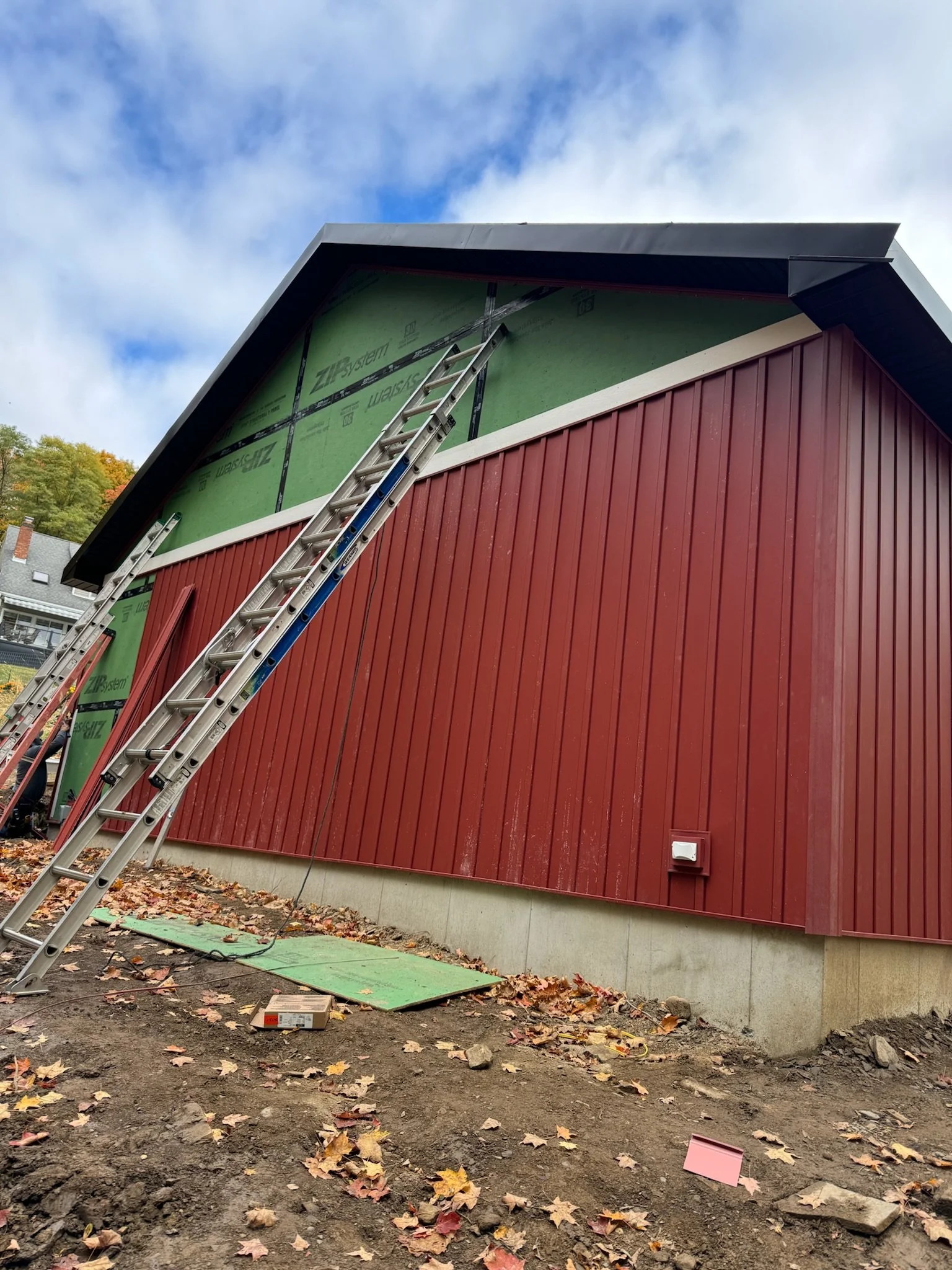 Side view of a house under construction with red exterior siding, green sheathing exposed at the upper section, and ladders leaning against it. Leaves are scattered on the ground.