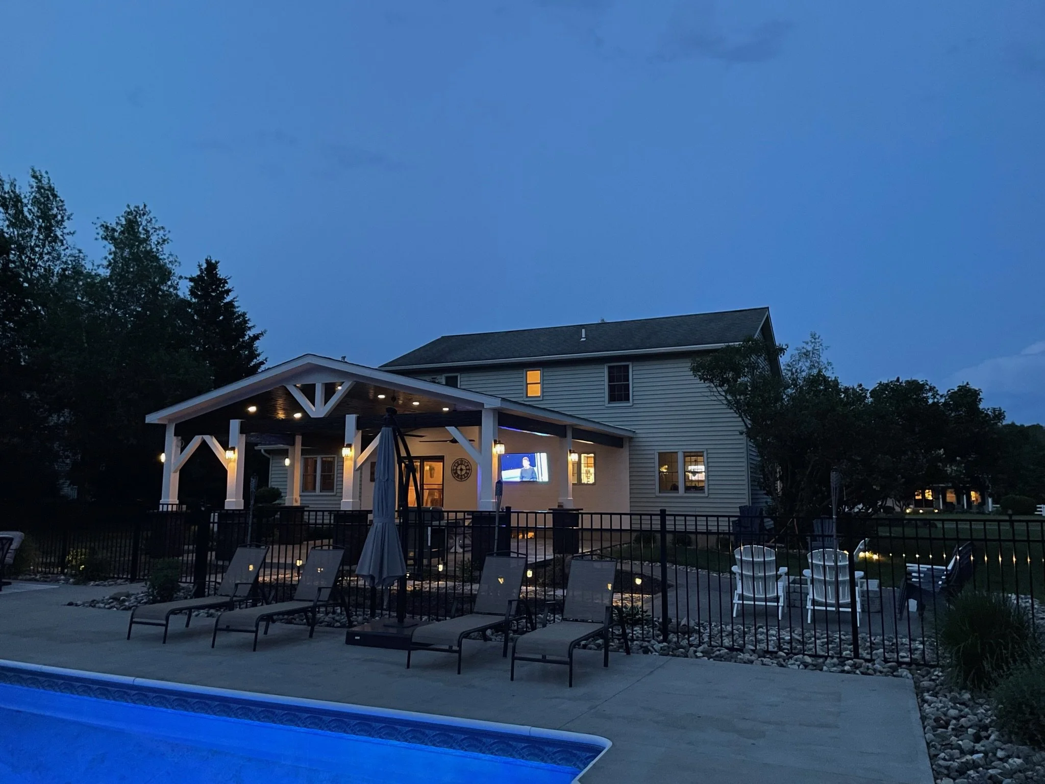 A backyard scene at dusk featuring a large two-story house with lit windows, an enclosed patio area with outdoor furniture including chairs and a table, a pool with a blue cover, and a fence surrounding the yard. The sky is darkening as evening sets