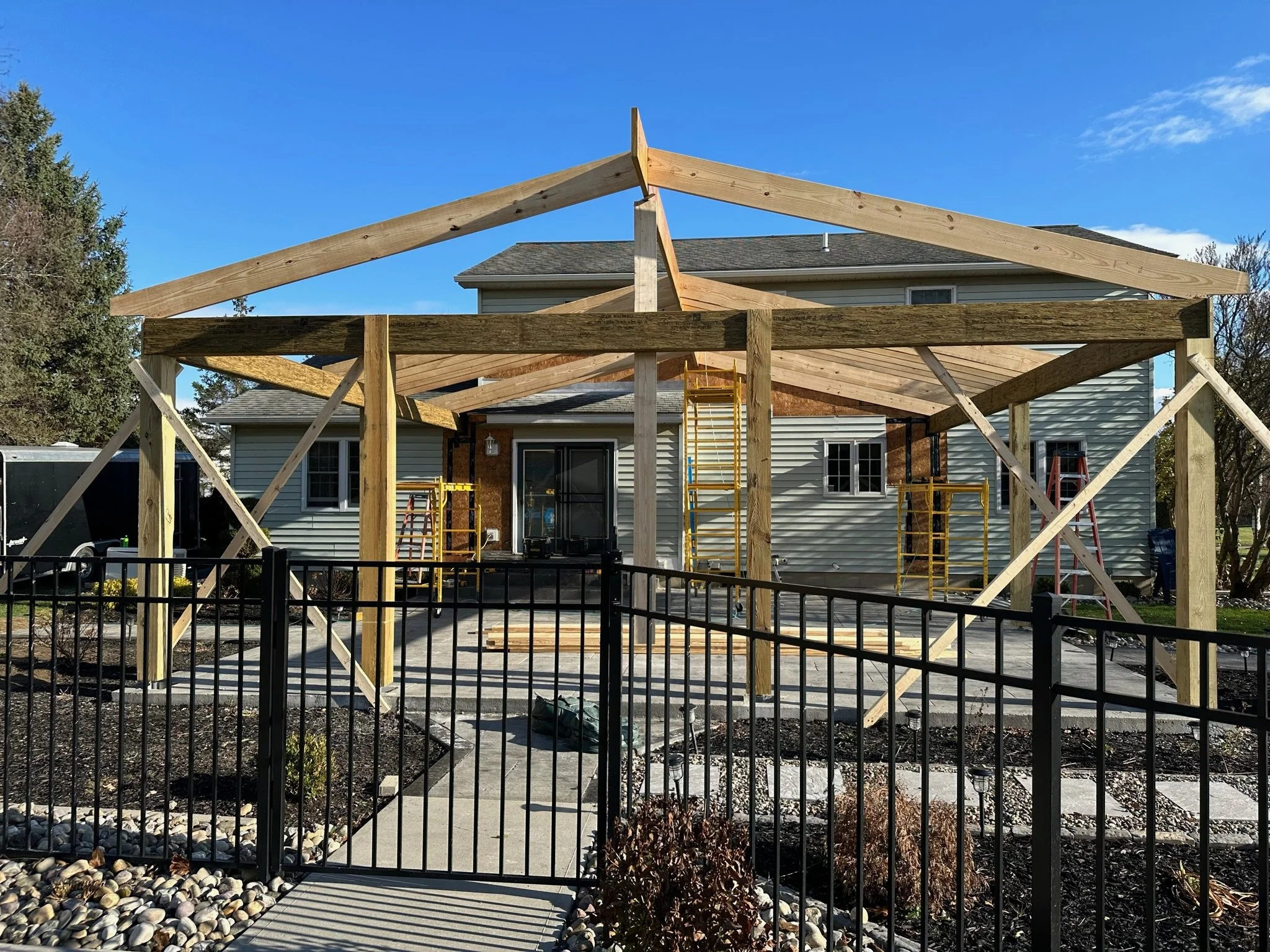 Construction of an outdoor patio or porch in progress in the backyard of a house, with wooden framing and support beams, tools, and scaffolding present.