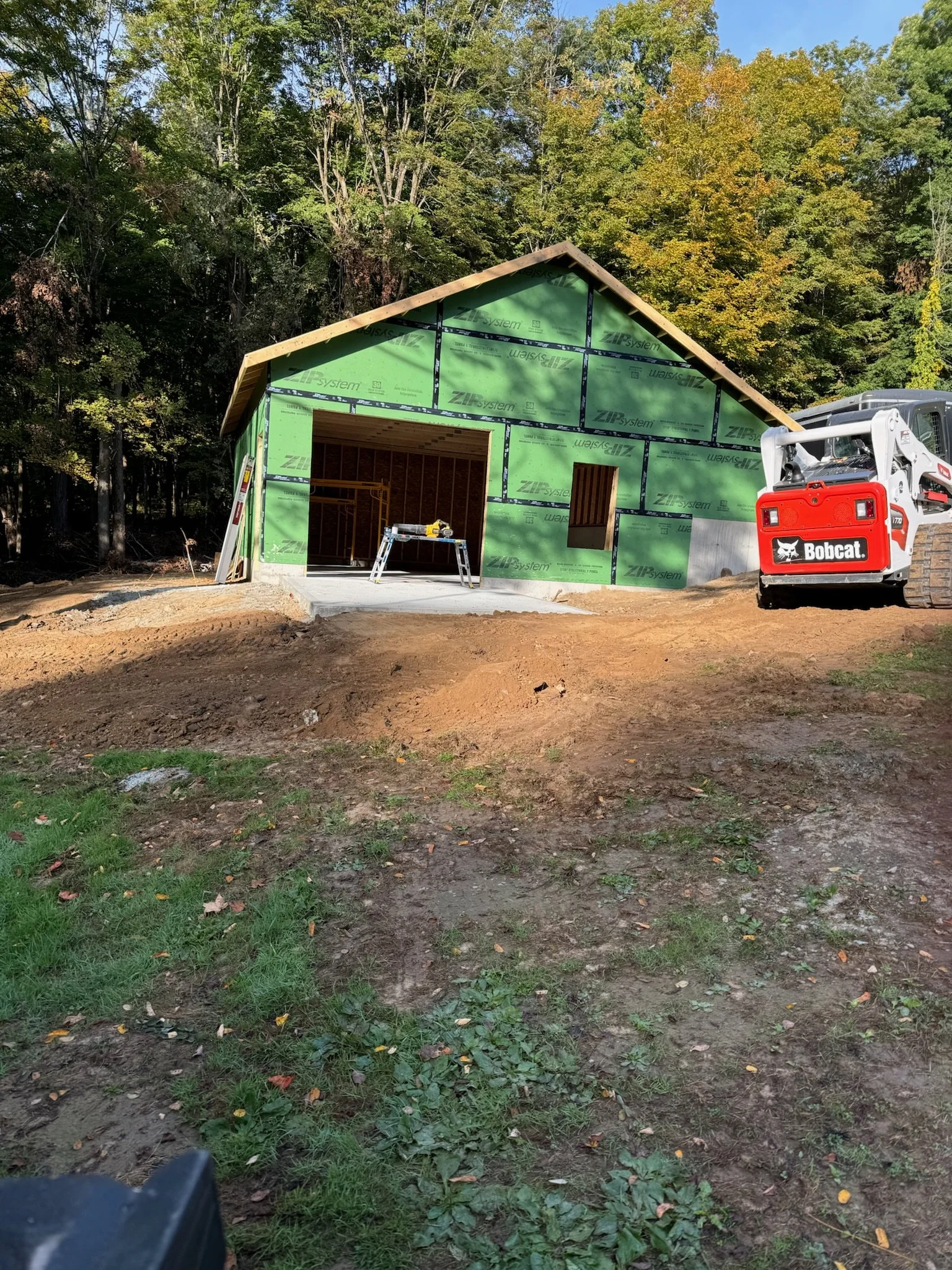 Construction site with a small house being built, green sheathing on the exterior walls, a wide opening for a garage door, a smaller window opening, a ladder, and construction equipment including a Bobcat loader.