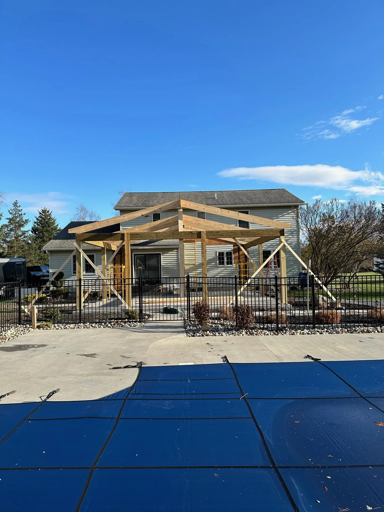 House under construction with a wooden frame structure, surrounded by a black metal fence, with a clear blue sky overhead.