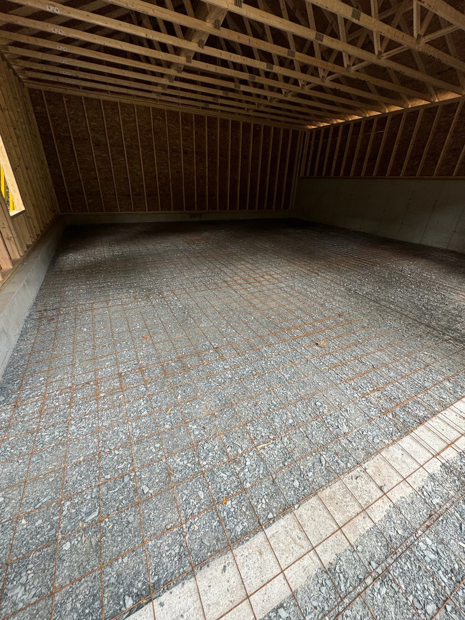 Interior of an unfinished attic with wooden framing, sloped roof, and gravel ground with a grid of rebar.