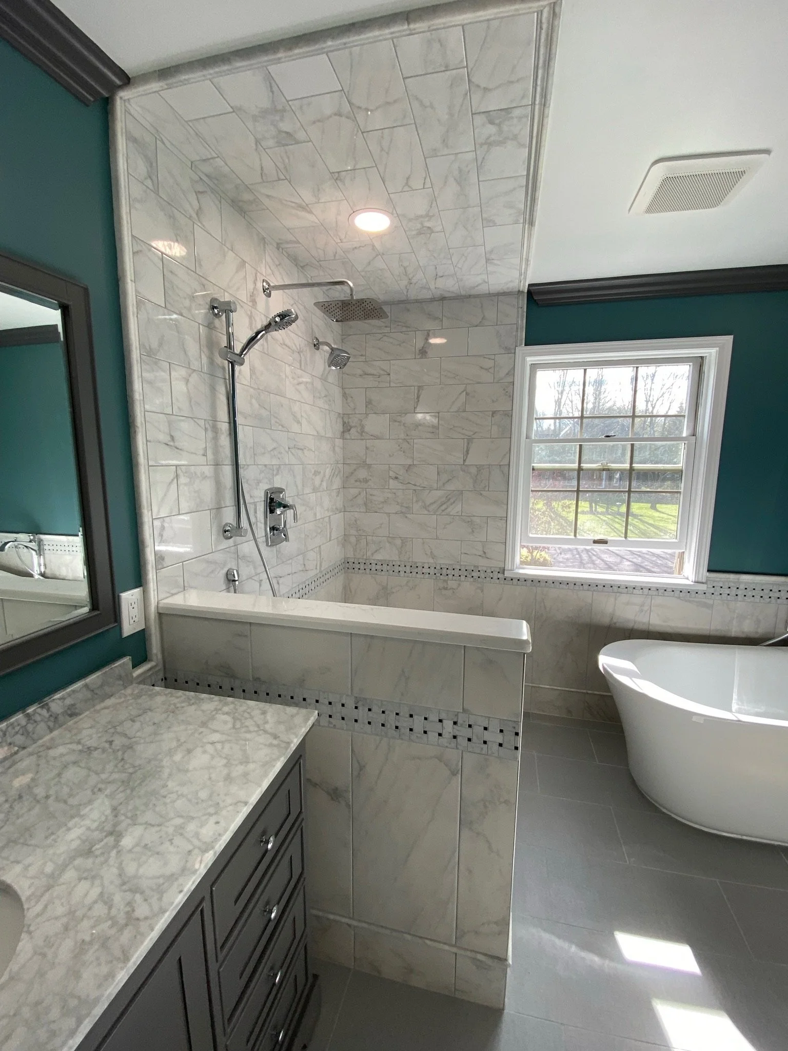 Modern bathroom with marble tiles, a glass-enclosed shower, a freestanding bathtub, and a window overlooking a green outdoor scene.