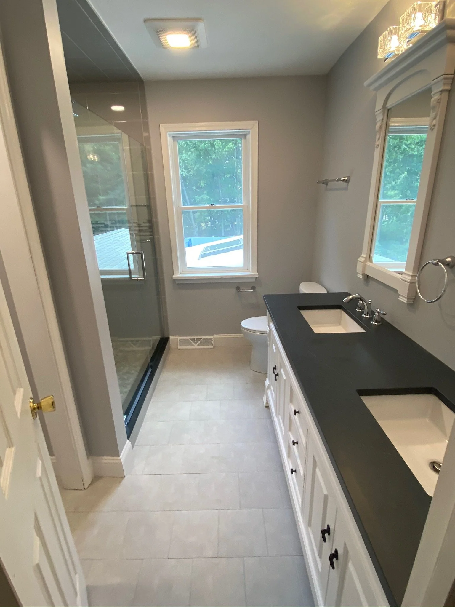 A bright bathroom with gray walls, a shower with glass doors, a white toilet, a black countertop with two integrated sinks, a mirror, and two windows showing greenery outside.
