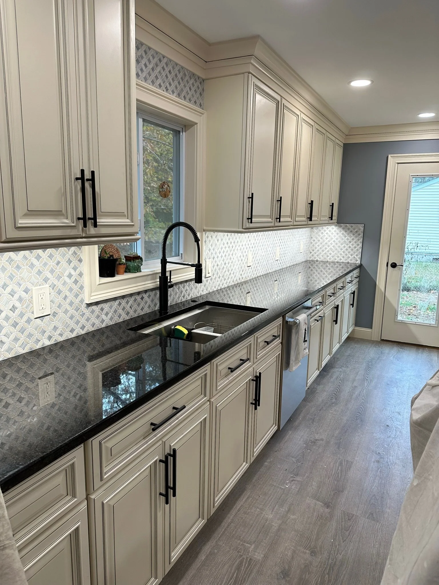 Kitchen with cream-colored cabinets, black handles, black granite countertop, and a patterned tile backsplash. There is a window above the sink with a potted plant on the window sill, and a door at the back leads outside. The flooring is wood-look. T