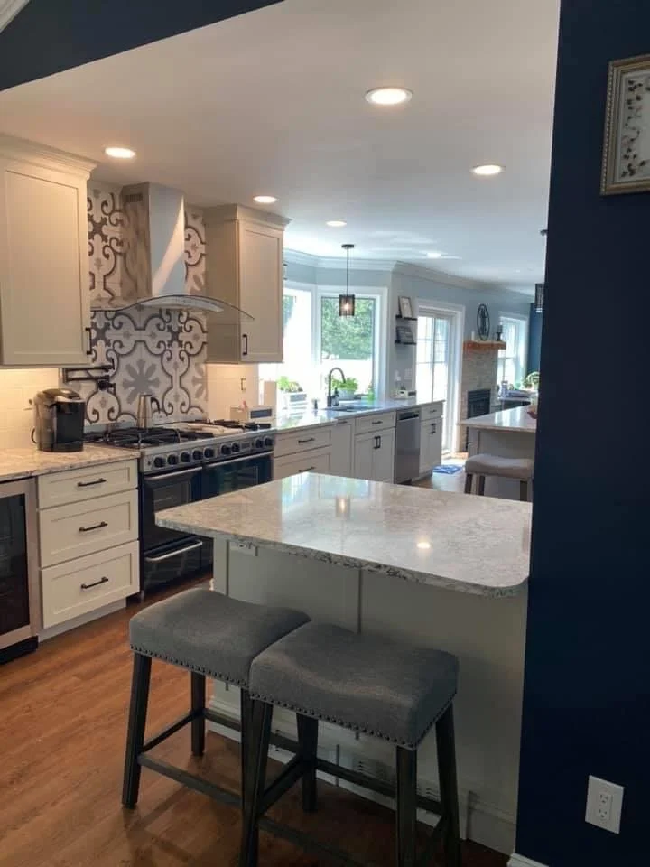 Modern kitchen with white cabinets, a marble countertop island, stainless steel appliances, and a patterned backsplash. There are two bar stools at the island, hardwood floors, and a view into a bright dining area with large windows.
