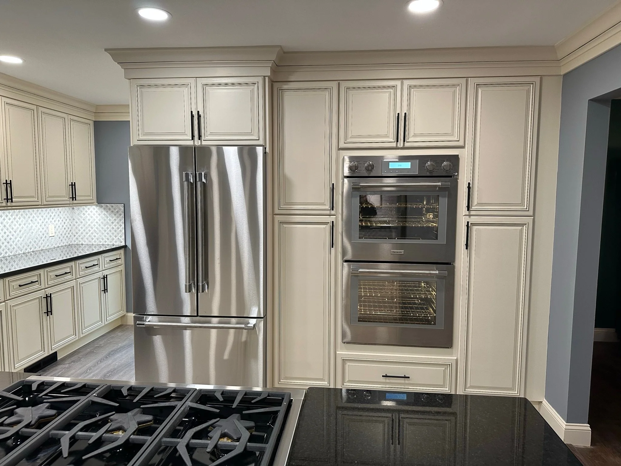 Modern kitchen with beige cabinetry, stainless steel refrigerator, double wall ovens, and black countertop with a gas stove in the foreground.