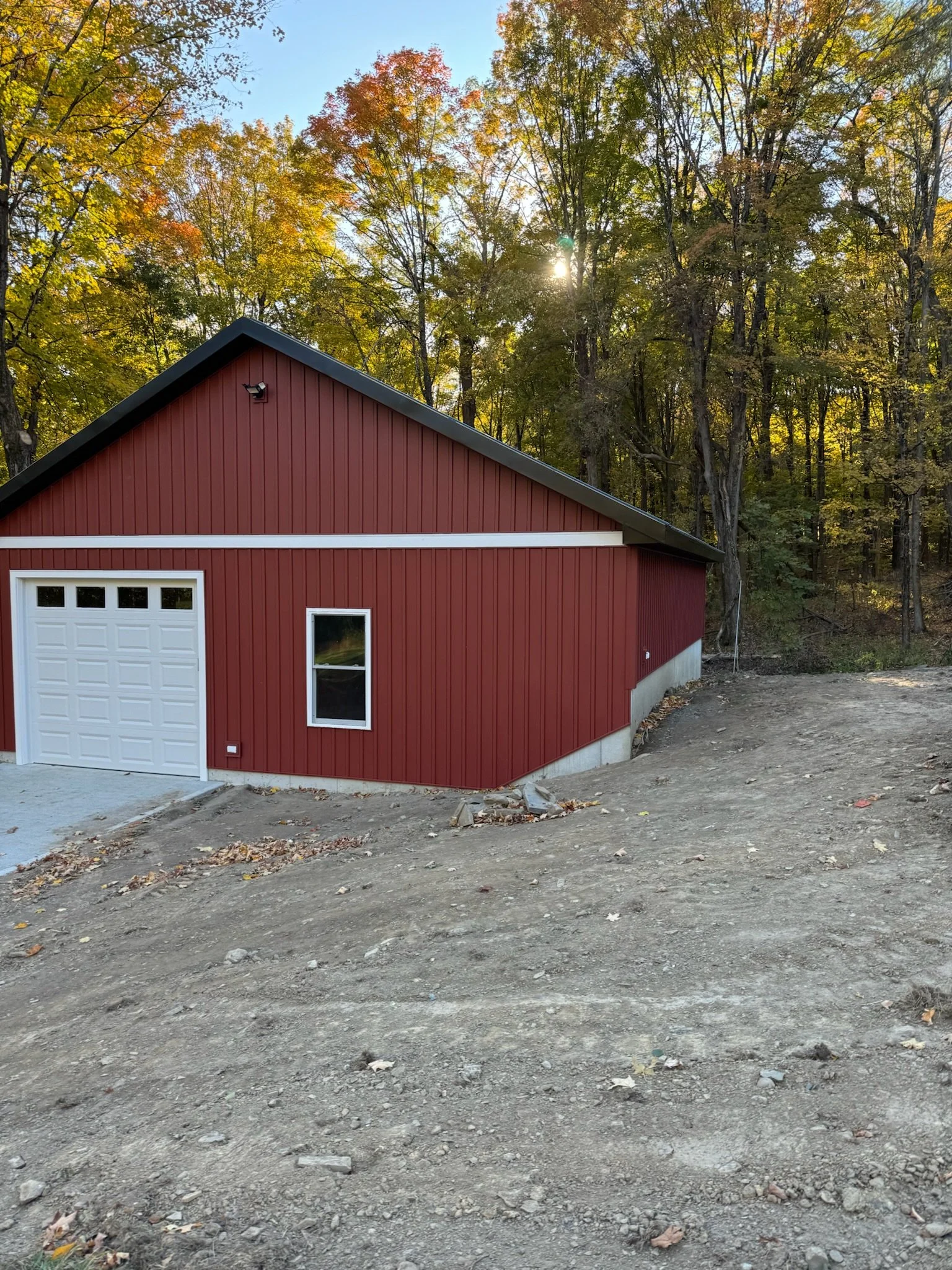 Red garage with white door and window, surrounded by an unfinished gravel driveway and a wooded area with fall-colored trees in the background.