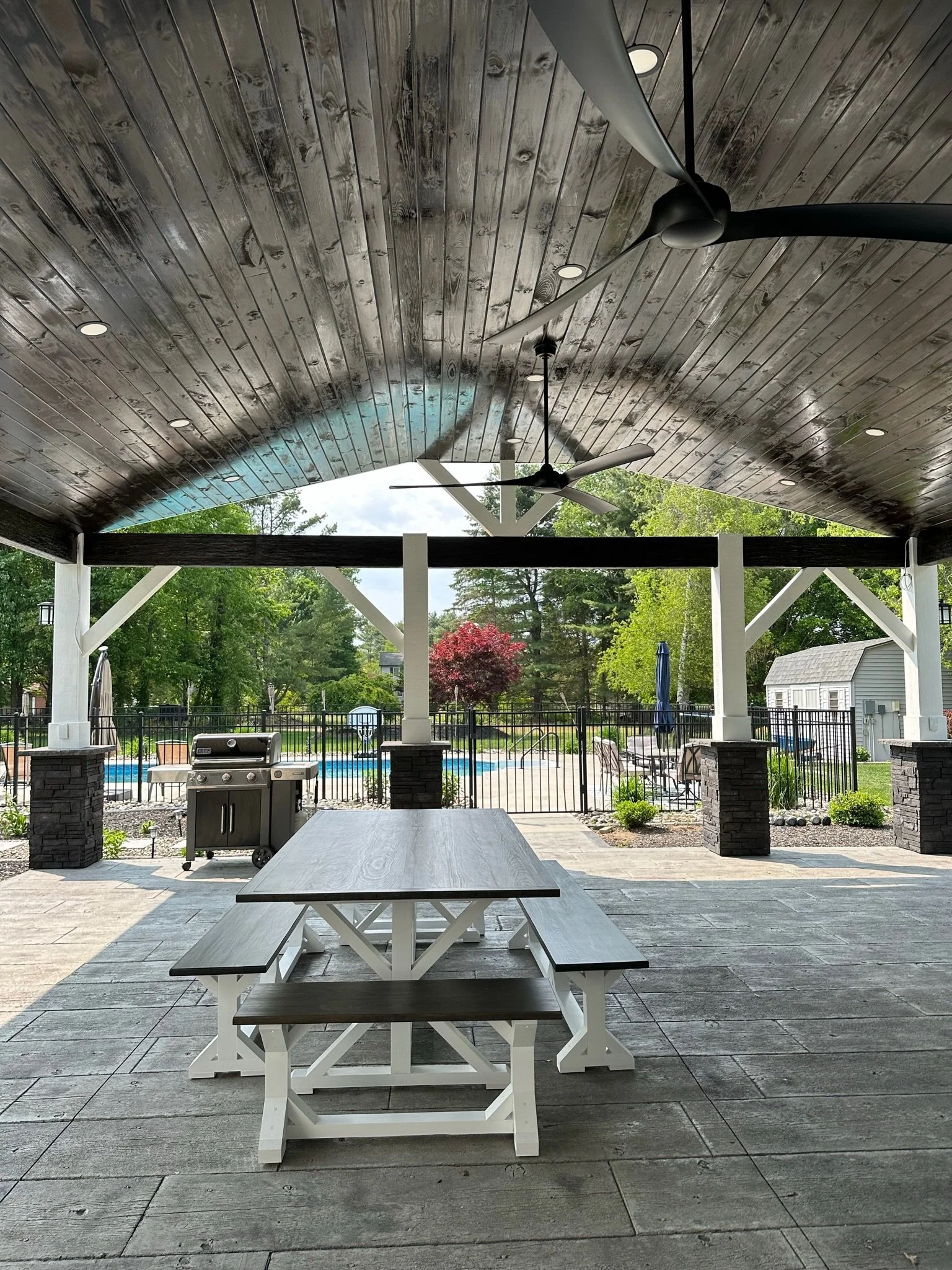 Covered outdoor patio area with a picnic table and bench, ceiling fans, a barbecue grill, and view of a swimming pool and yard with trees and plants.