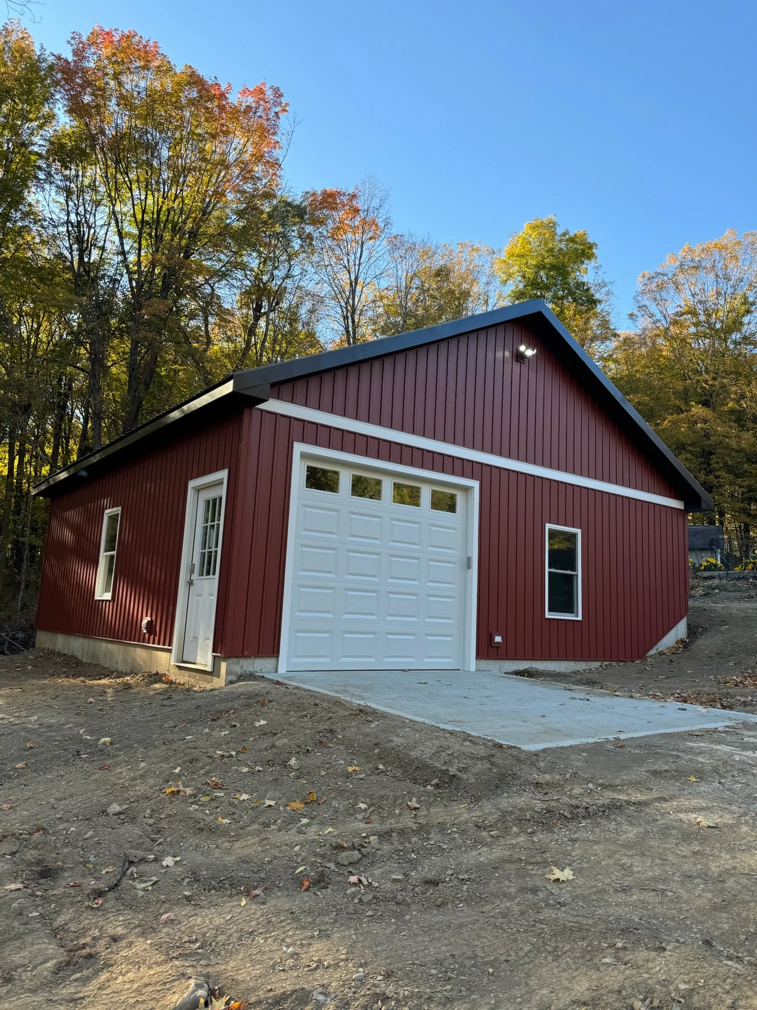 Red garage building with white door and trim, situated on a concrete driveway with cleared dirt yard, surrounded by trees with fall foliage, under a clear blue sky.