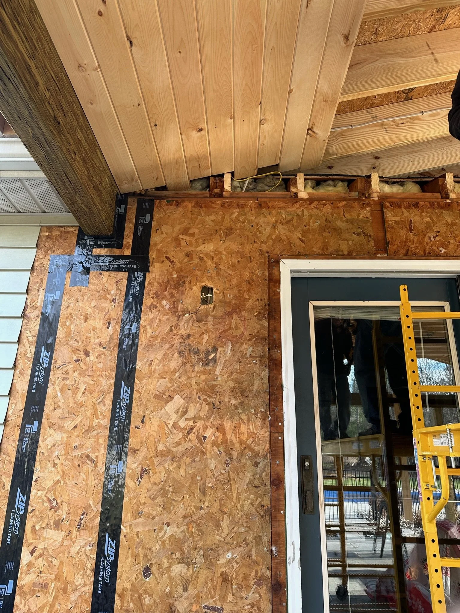 Part of a house under construction or renovation, showing wood paneling and insulation, with a ladder and black flashing tape on the wall near a sliding door.