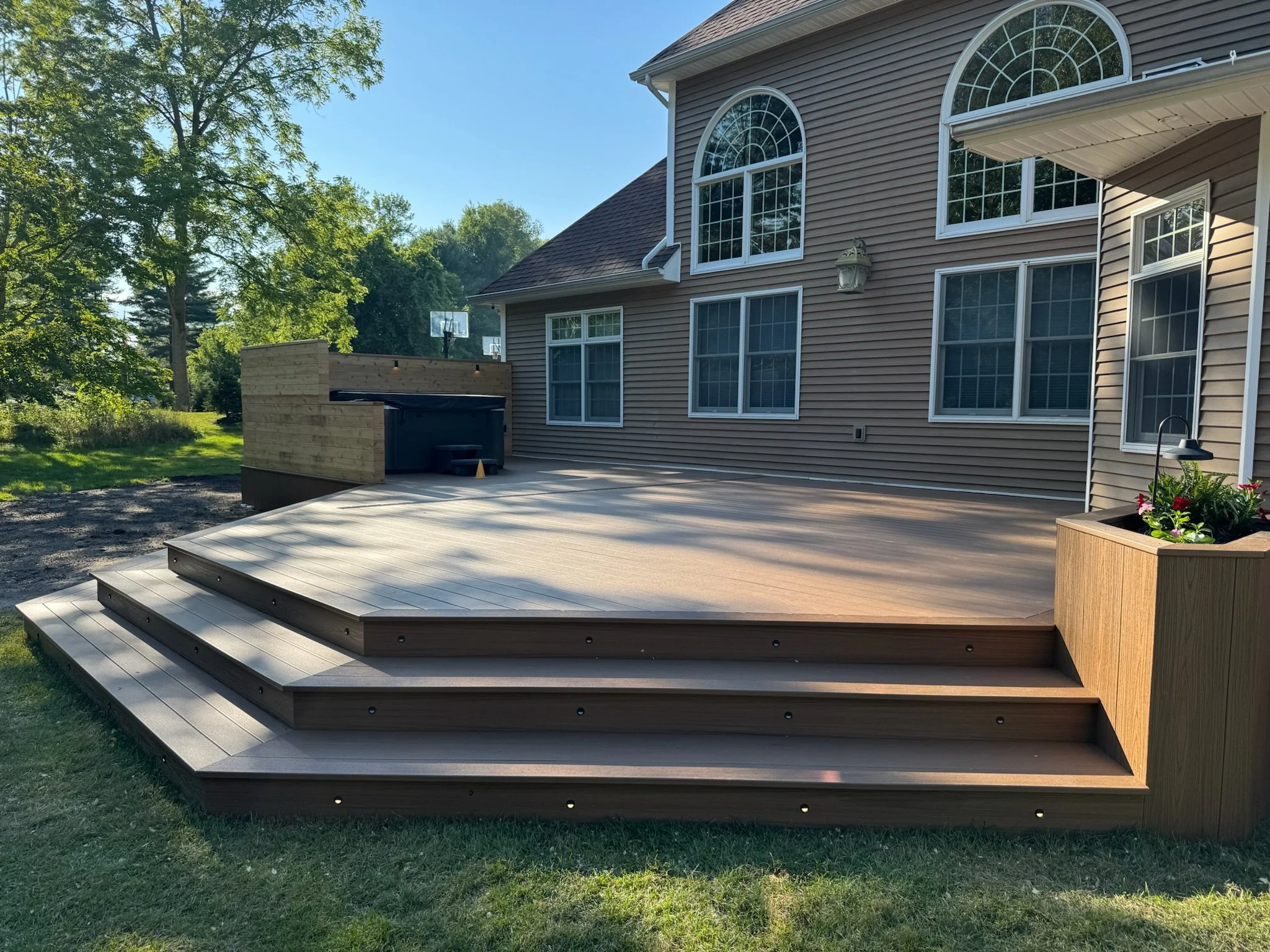 A newly built wooden deck attached to a house in a backyard, featuring steps leading to a grassy area and a planter with flowers on the corner.