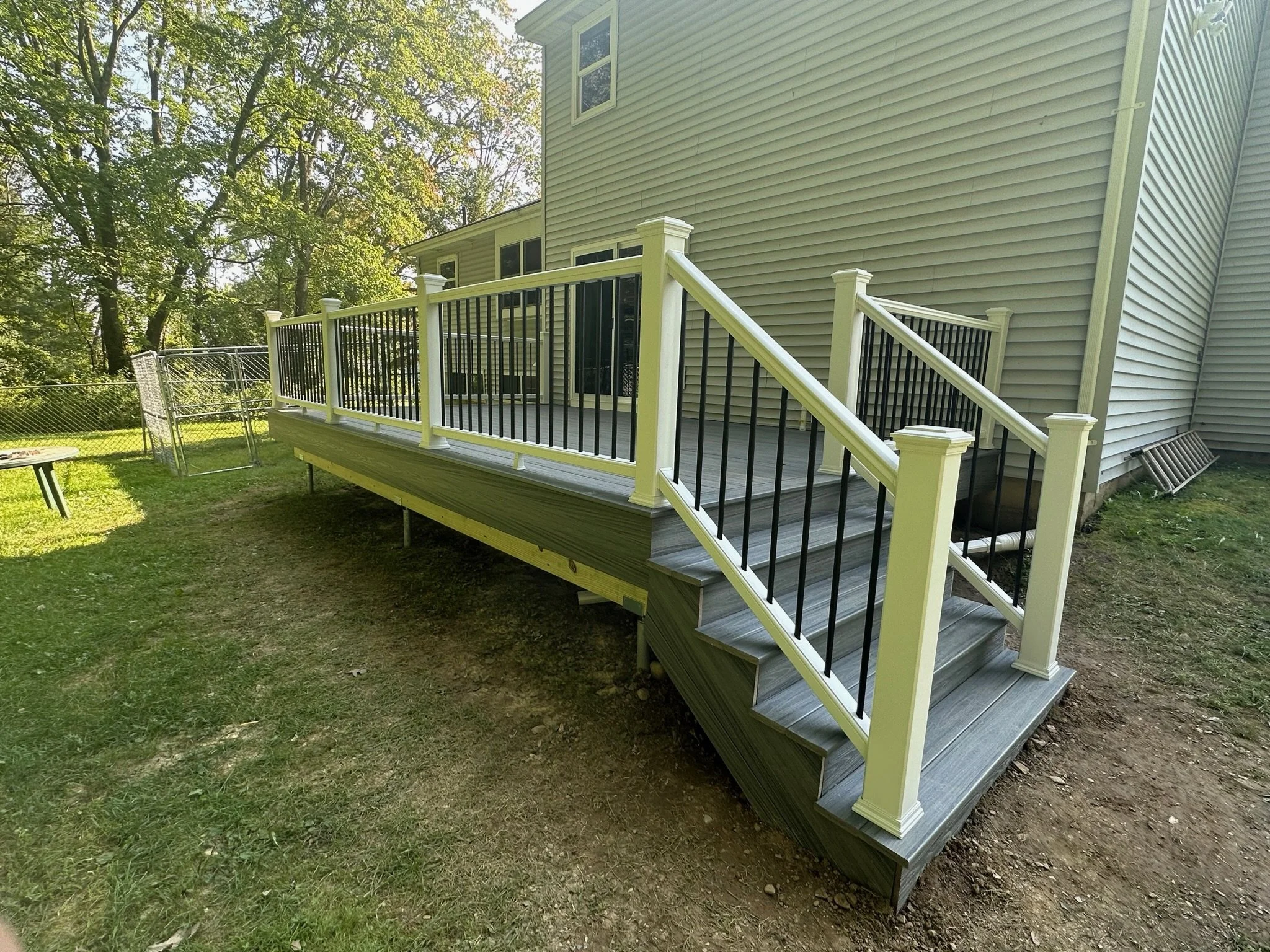 New wooden deck with black and white railing attached to the back of a house, overlooking a grassy yard with trees.