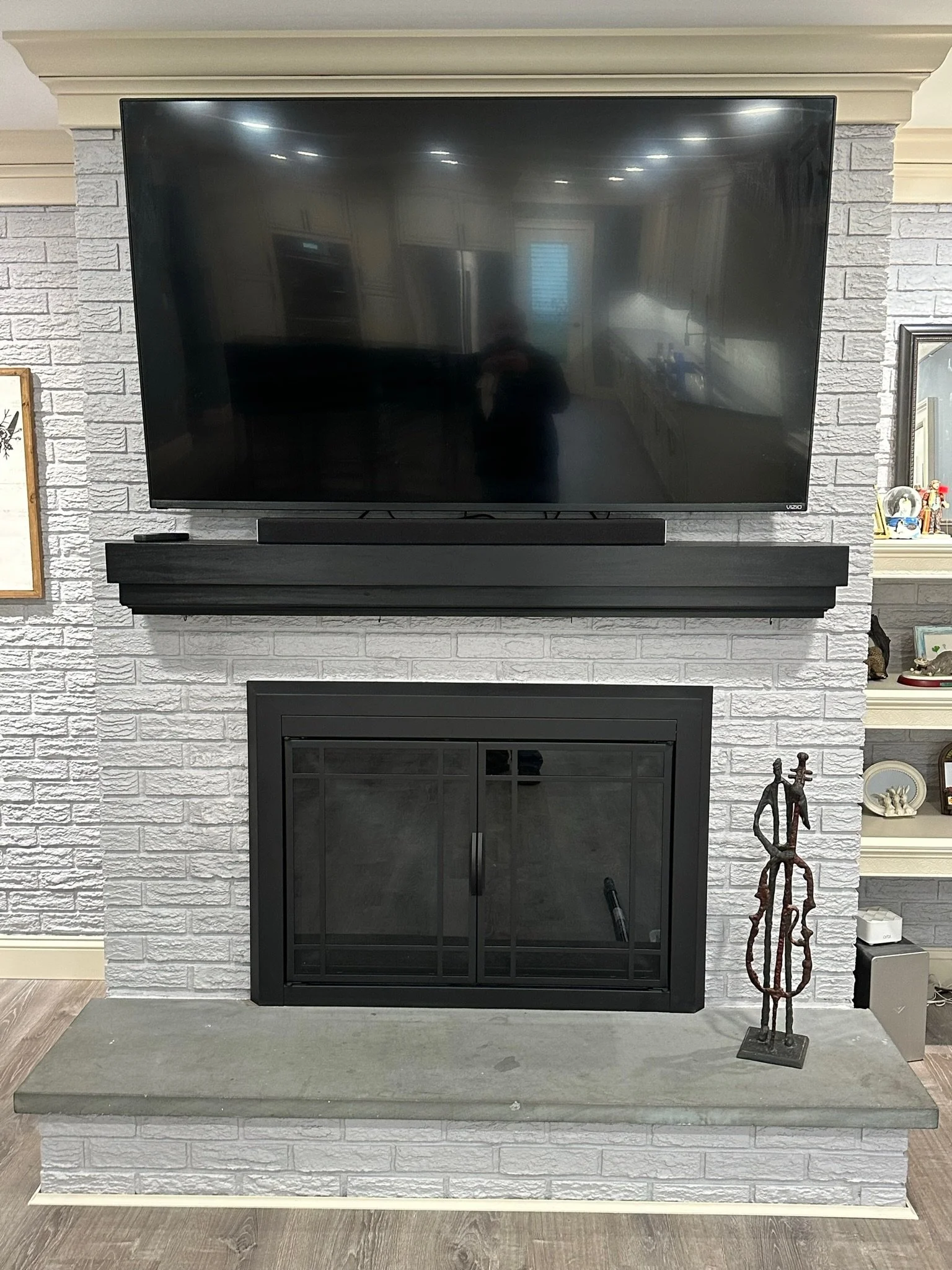 A living room fireplace with a mounted flat-screen TV above it. The fireplace is surrounded by white brick and has a gray stone hearth. To the right, there is a sculpture on the hearth and a built-in white shelf with decorative items.