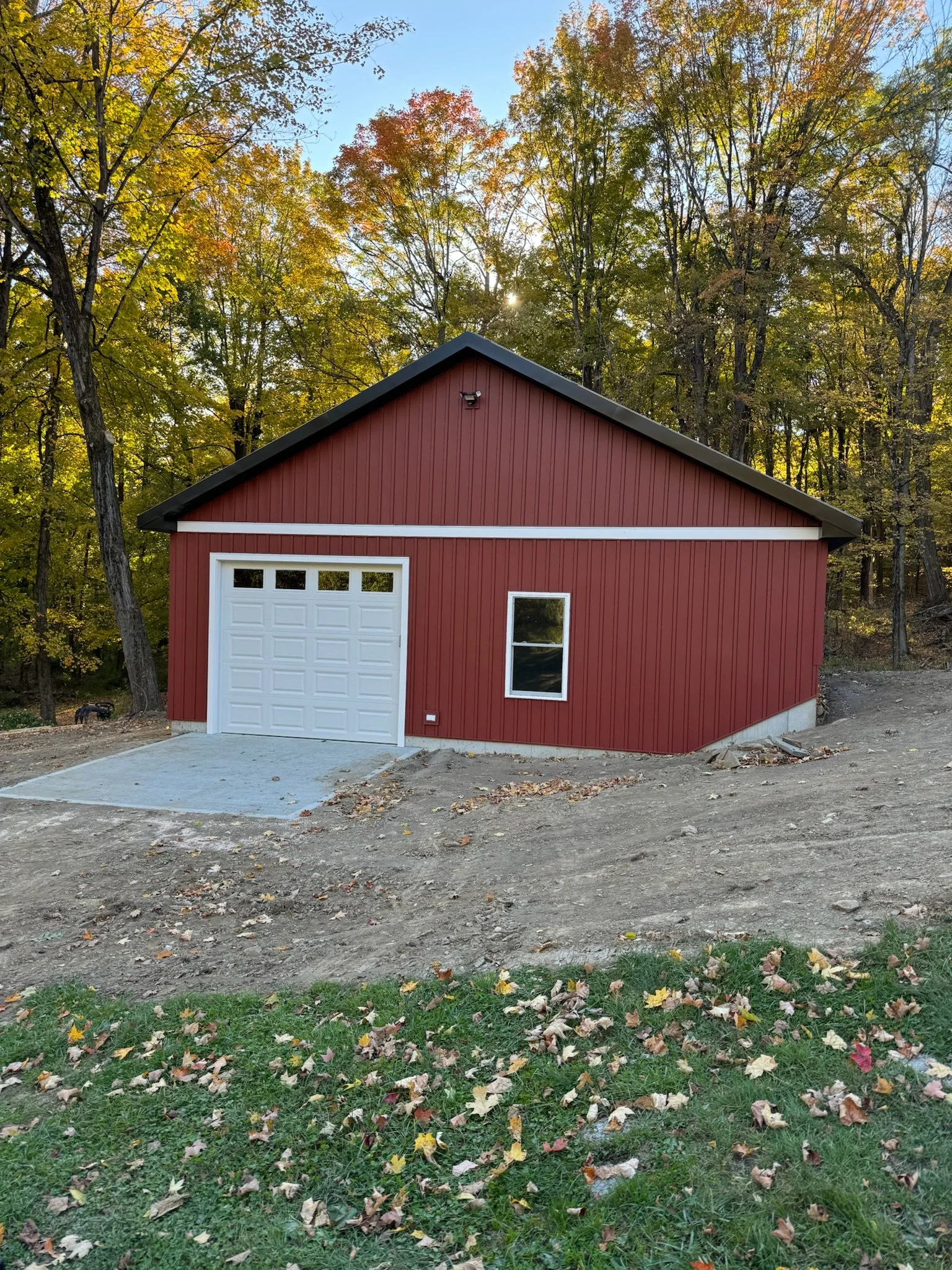 A red garage with a white door and a small window, surrounded by a dirt yard and trees with autumn leaves.