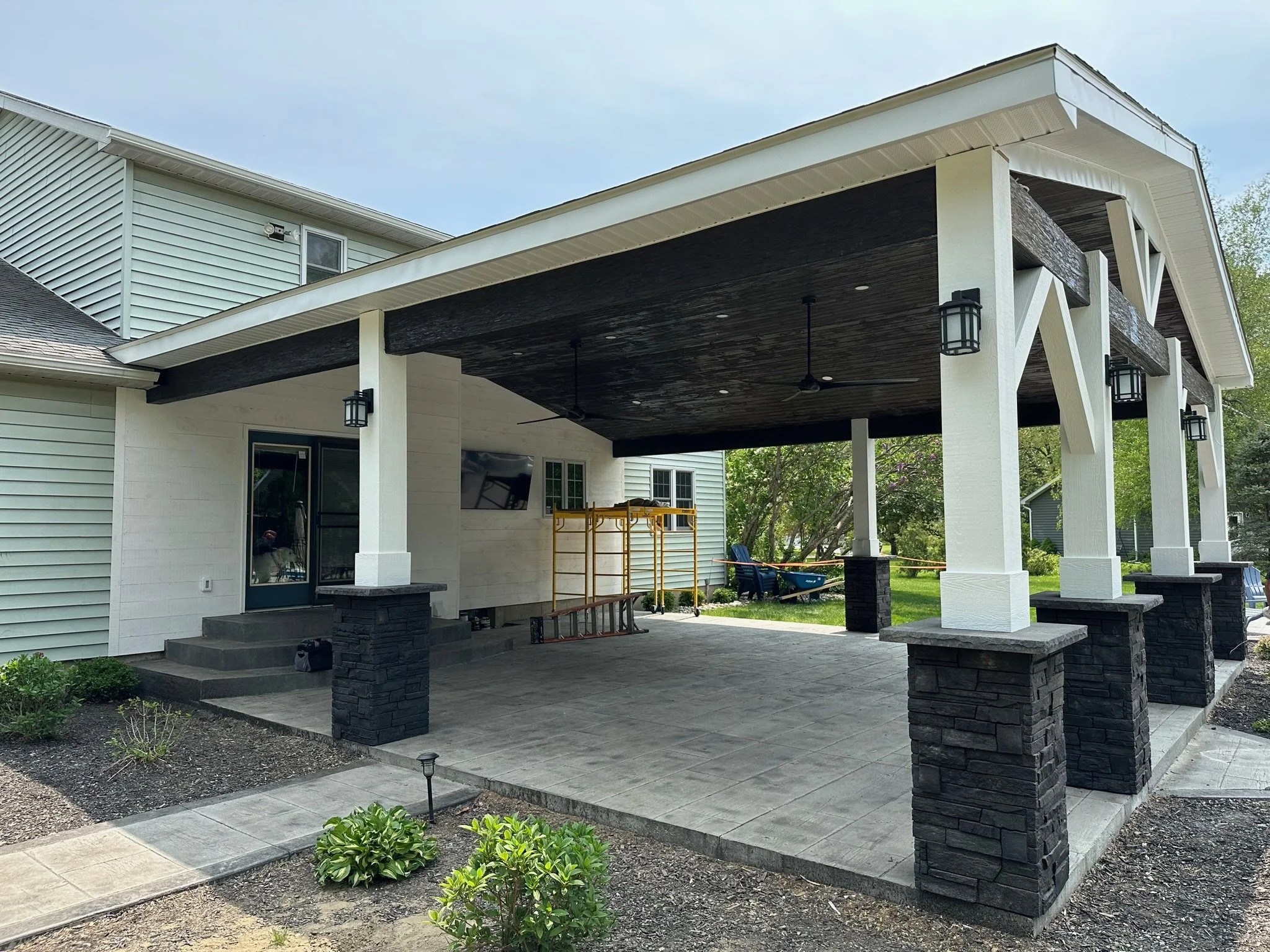 Backyard patio with a covered area featuring black and white columns, ceiling fans, mounted TV, and outdoor lighting, with garden and neighboring house in the background.