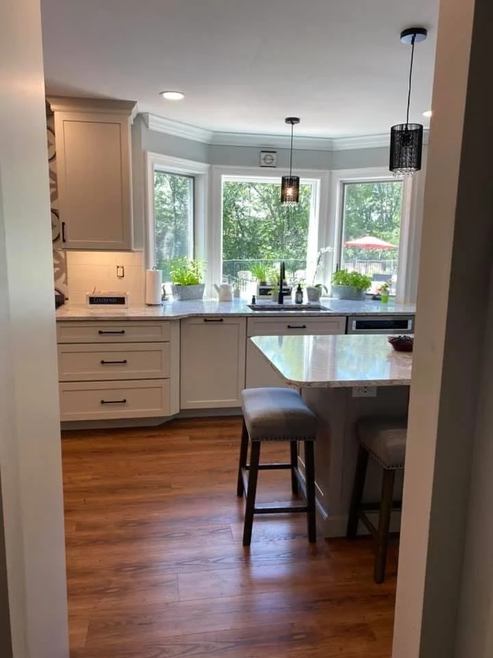 Bright kitchen with white cabinets, a marble countertop, and large bay windows with greenery outside. There are two hanging pendant lights, a small kitchen island with seating, and some plants on the windowsill.