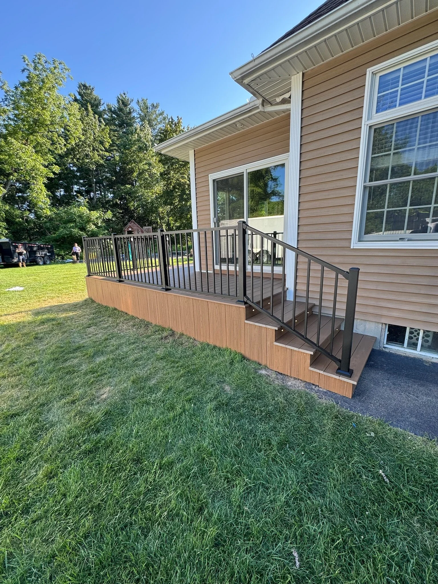 A wooden deck with black metal railing attached to the back of a house with beige vinyl siding and white trim. The deck has stairs leading down to a grassy yard.