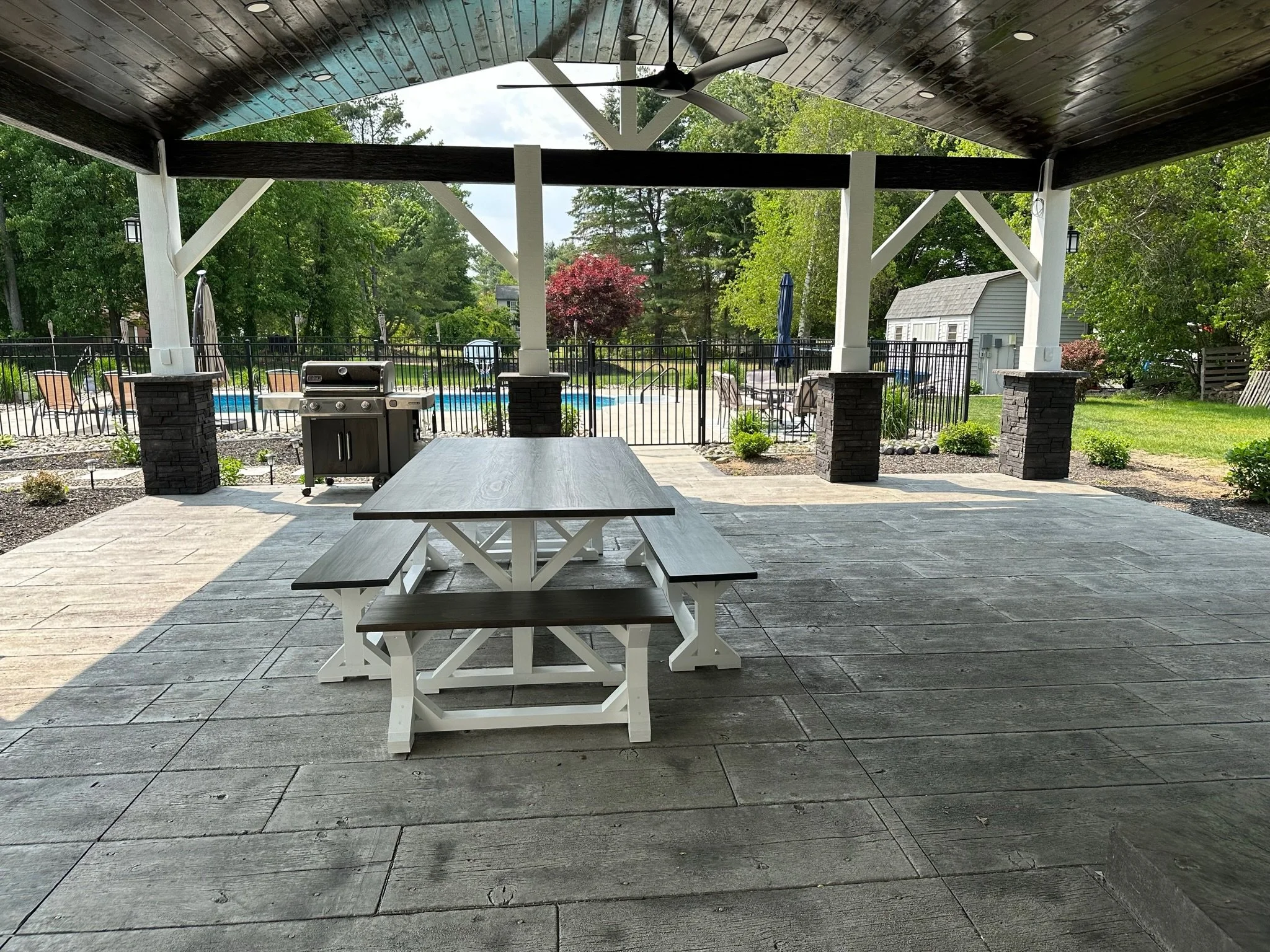 Covered patio with a picnic table, grill, and ceiling fan, overlooking a backyard with a swimming pool, lounge chairs, and trees.