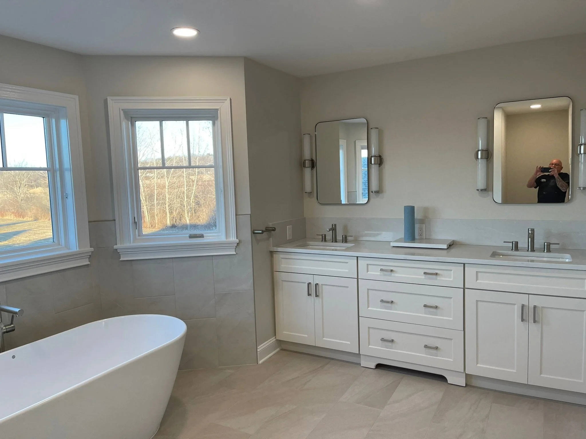 Modern bathroom with white vanity cabinets, two mirrors, two sinks, and a freestanding bathtub by a window in a bright room.