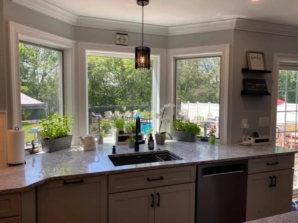 Kitchen with large windows overlooking a backyard, white cabinets, a marble countertop with plants and kitchen accessories, and a black pendant light hanging from the ceiling.