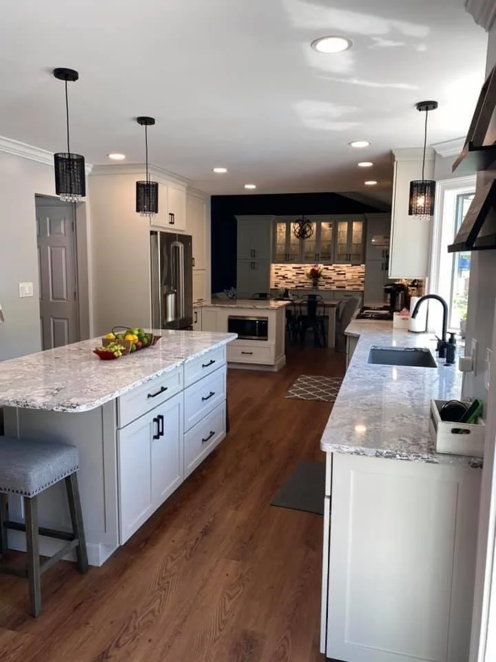 Modern kitchen with white cabinets, granite countertops, stainless steel refrigerator, pendant lighting, hardwood floors, and a dining area in the back.