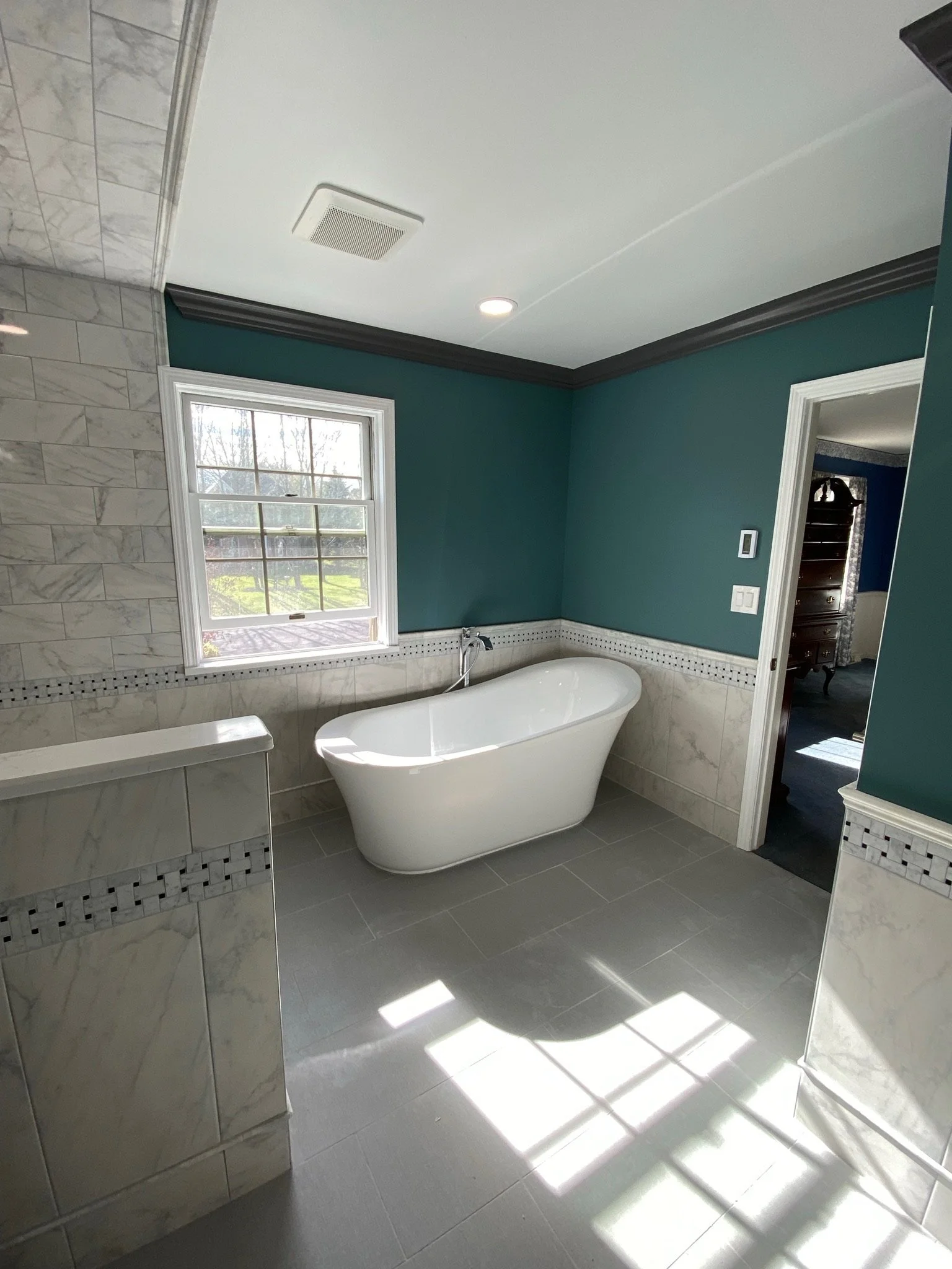 A bathroom with a white standalone bathtub beneath a window, teal walls, marble tiles, and gray floor tiles. Sunlight streams through the window, creating patterns on the floor.