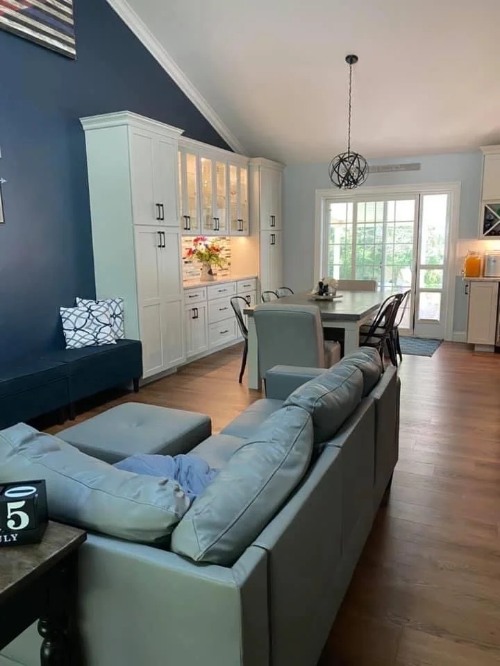 Living room with white sectional sofa, blue and white pillows, and a blue accent wall behind a white built-in cabinet. Dining table with chairs and a spherical pendant light are visible. Large window with a door leading outside, hardwood floors throu