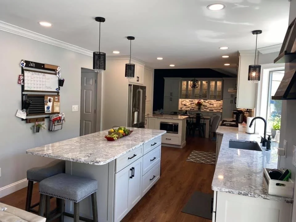 Modern kitchen with white cabinets and granite countertops, an island with stools, and a breakfast nook in the background. Pendant lights hang above the island, and a wall-mounted calendar and organizer are on the left wall. There is a stainless stee