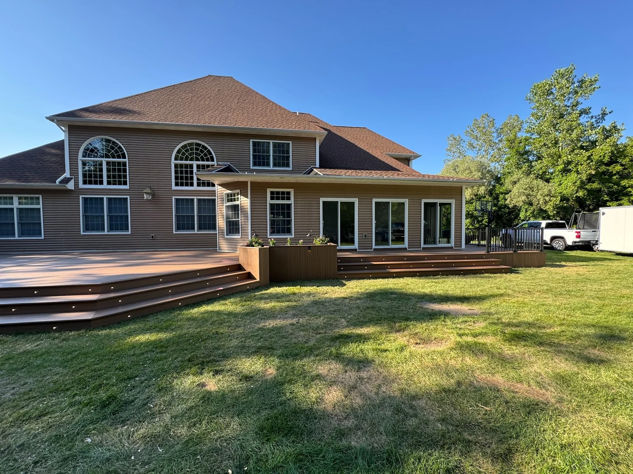 View of a two-story house with a large wooden deck, sliding glass doors, and multiple windows in a grassy backyard, with trees and parked cars in the background.