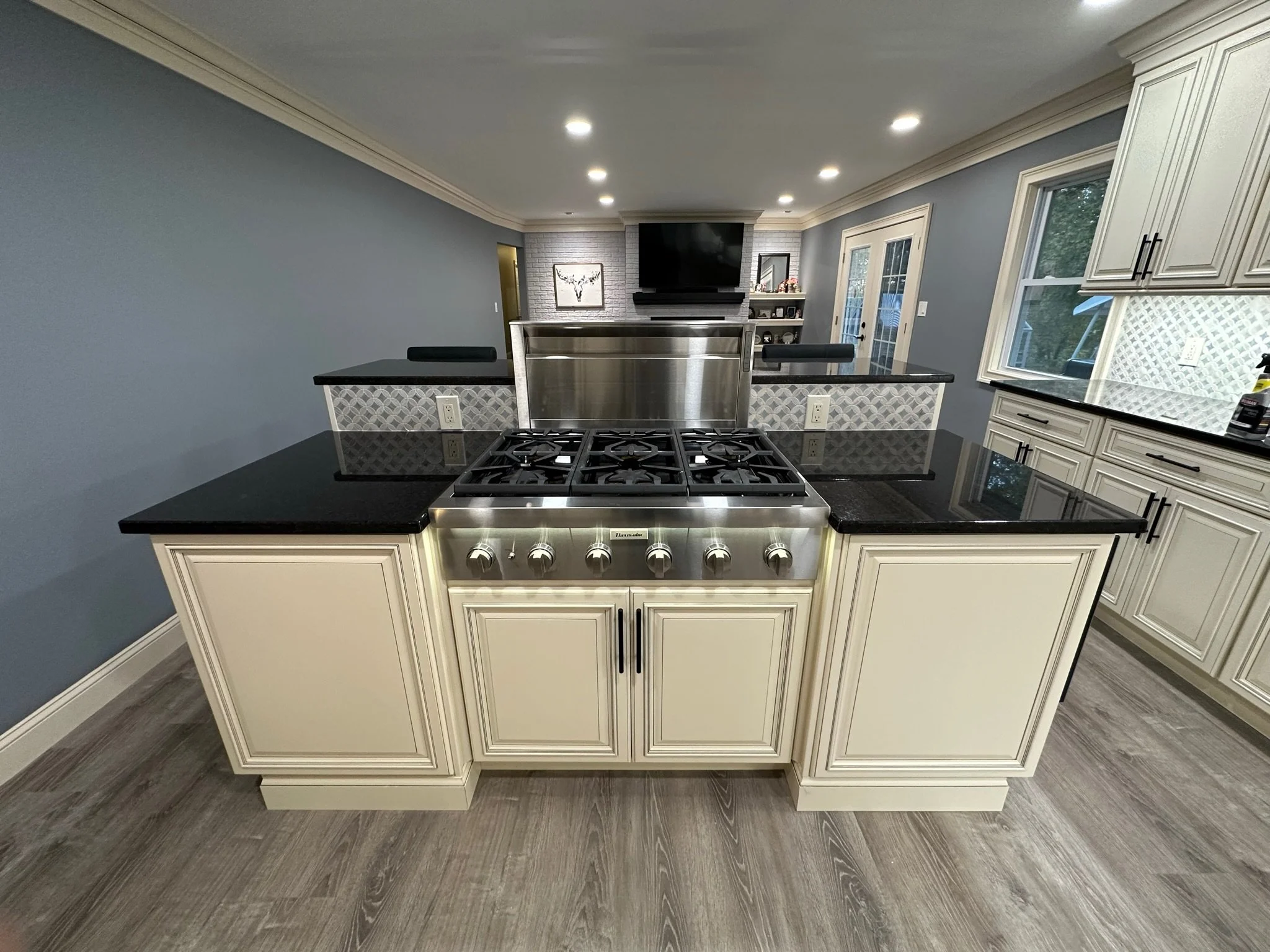 Kitchen with black countertops, cream cabinetry, a stainless steel gas stove with six burners, a silver backsplash, and a view of a living room with TV and shelves.