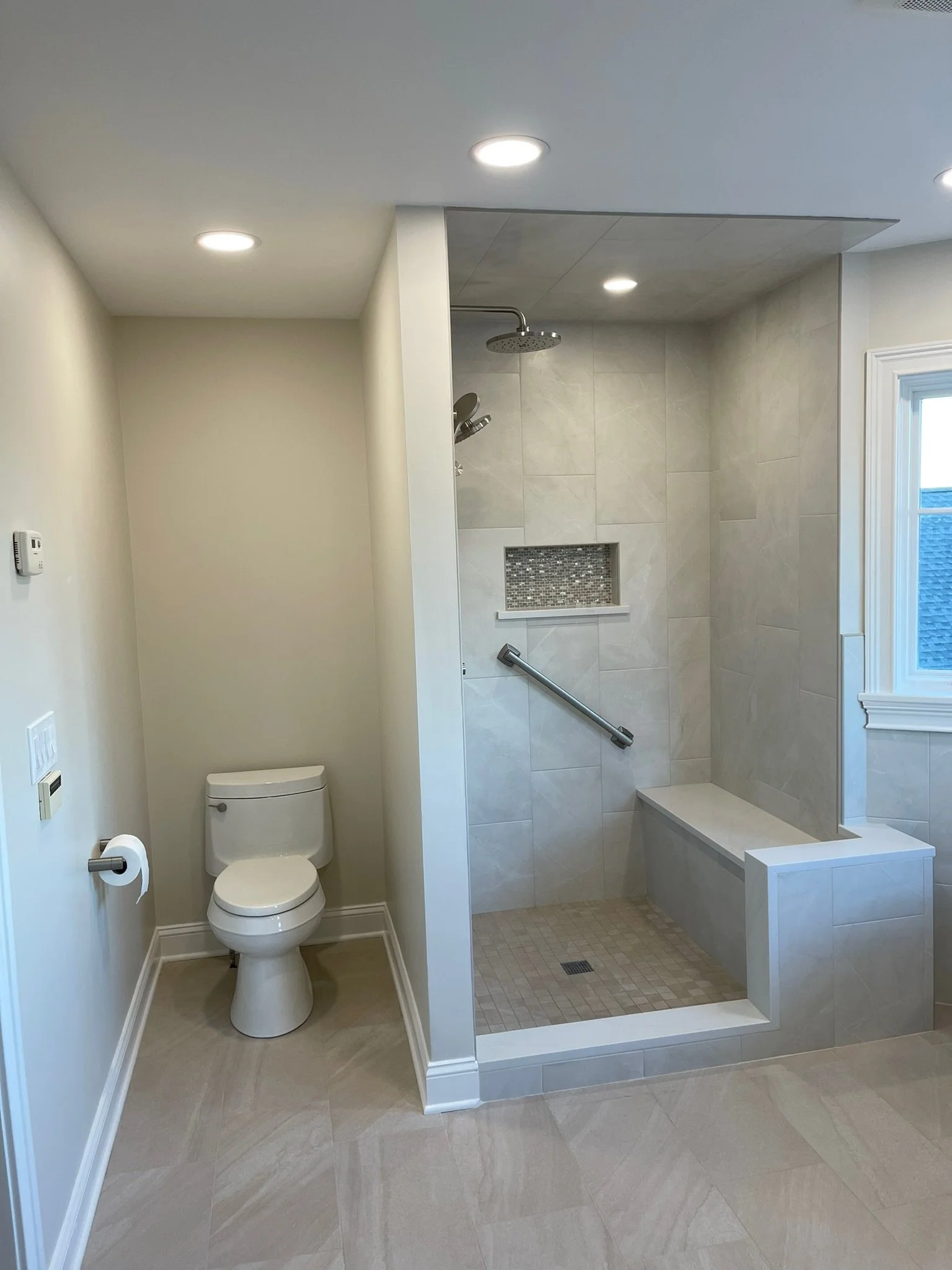 Bathroom with a toilet on the left and a walk-in shower on the right, featuring a showerhead, a built-in shelf with mosaic tiles, and a bench, with a large window on the far right.