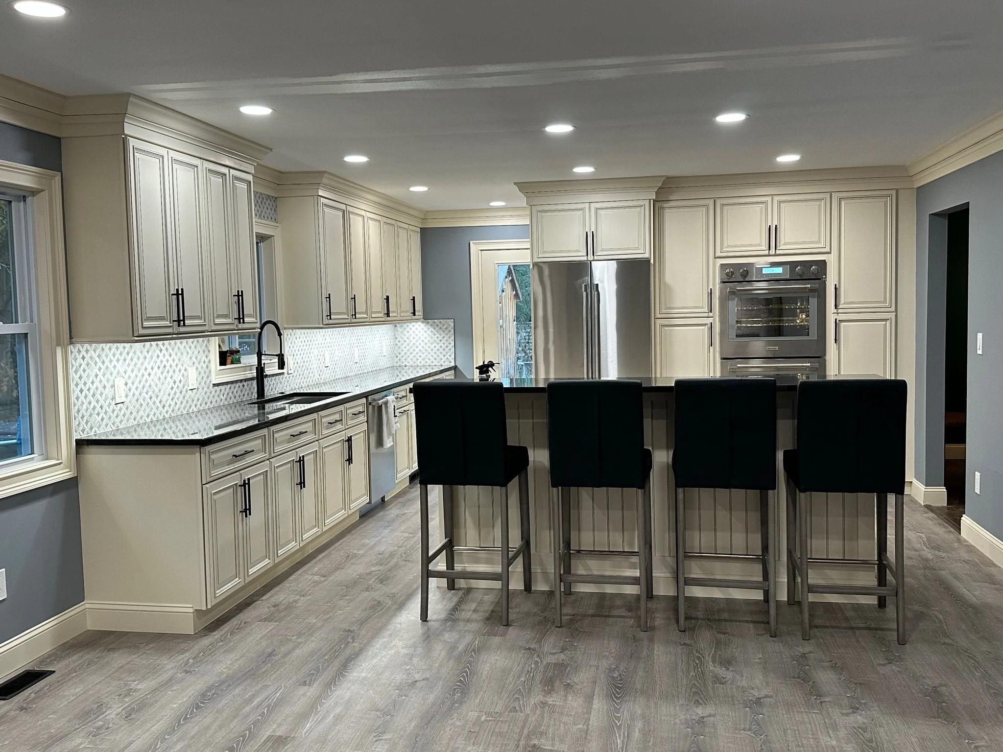 Modern kitchen with white cabinetry, black countertops, a central island with four black barstools, stainless steel appliances, and a light wood floor.