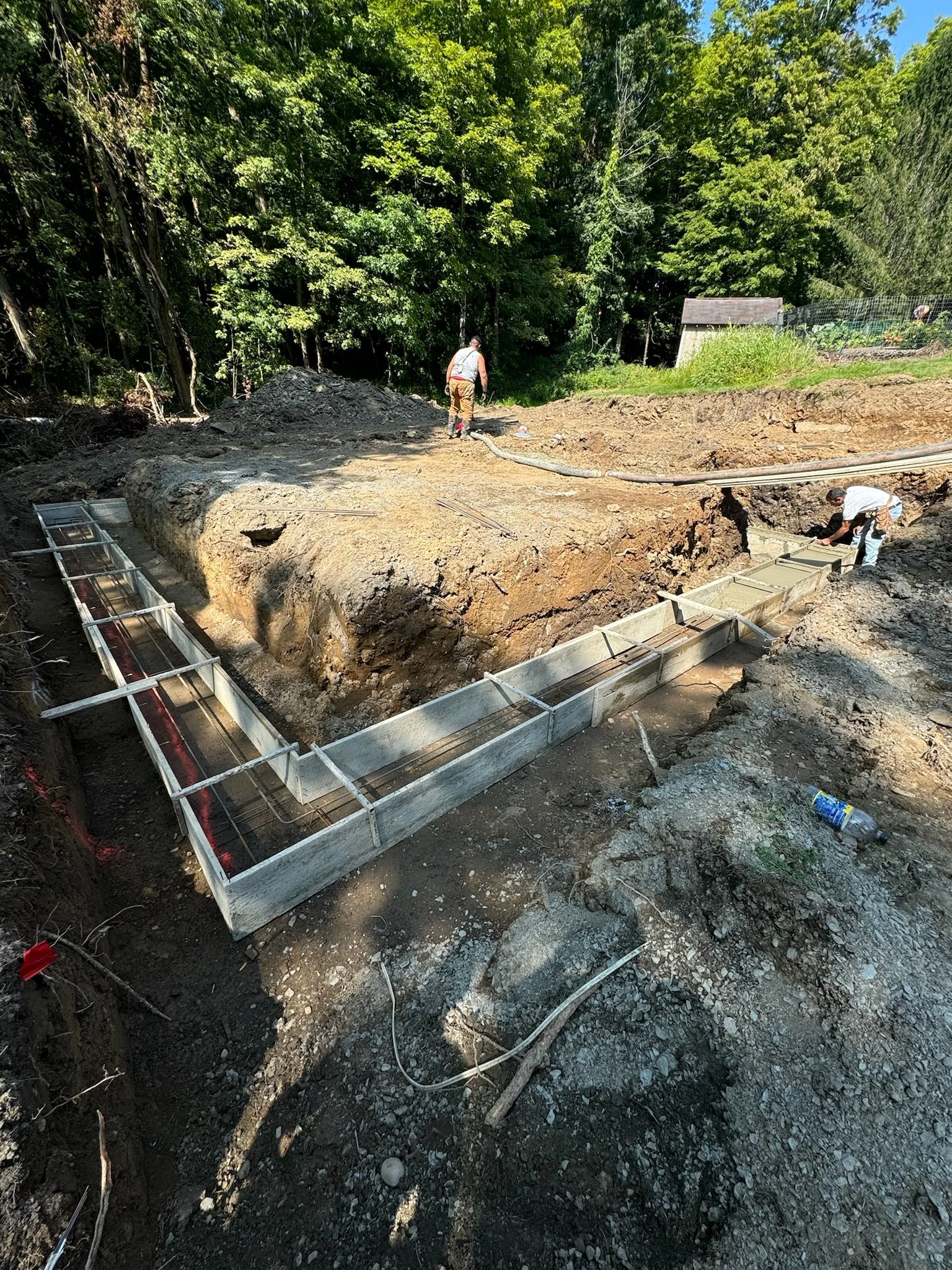 Construction site with metal formwork for a concrete foundation being prepared, workers working, dirt excavation, and wooded background.