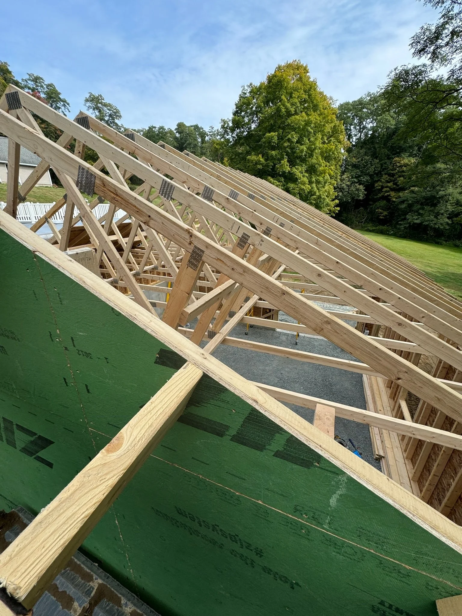 Construction site with wooden roof trusses and green exterior wall sheathing under a partly cloudy sky, trees in the background.