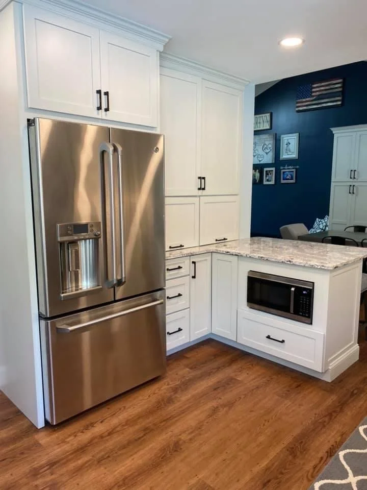 A modern kitchen corner featuring a stainless steel refrigerator with a water and ice dispenser, white cabinetry with black handles, a granite countertop, a built-in microwave, and a hardwood floor.