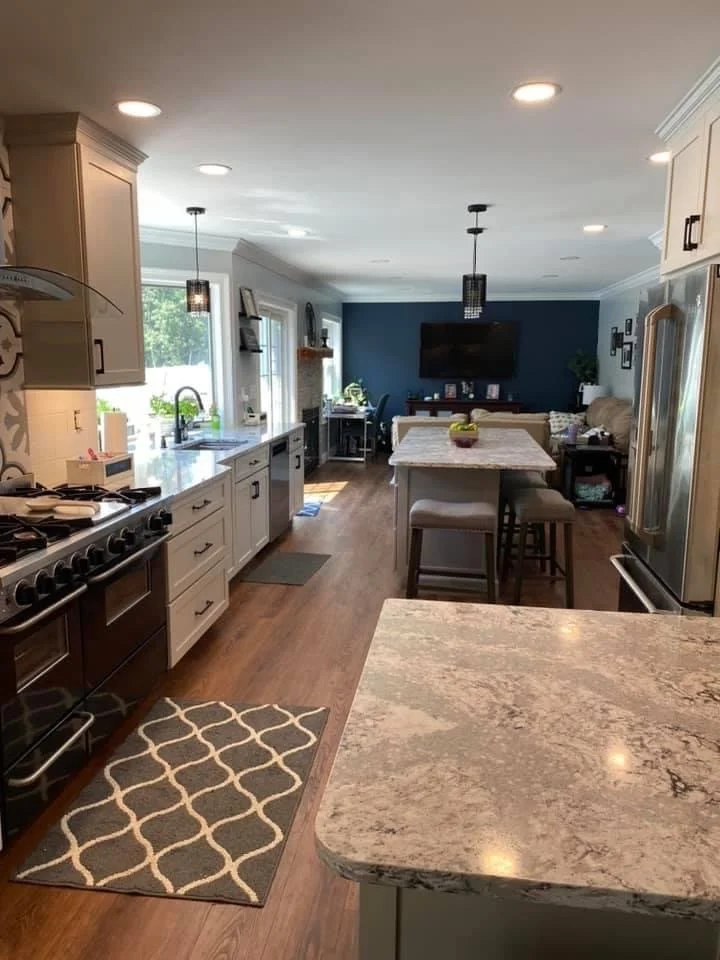 A spacious kitchen with white cabinetry, a large marble island, stainless steel appliances, and a dark blue accent wall in the living area with a mounted TV and sofa.