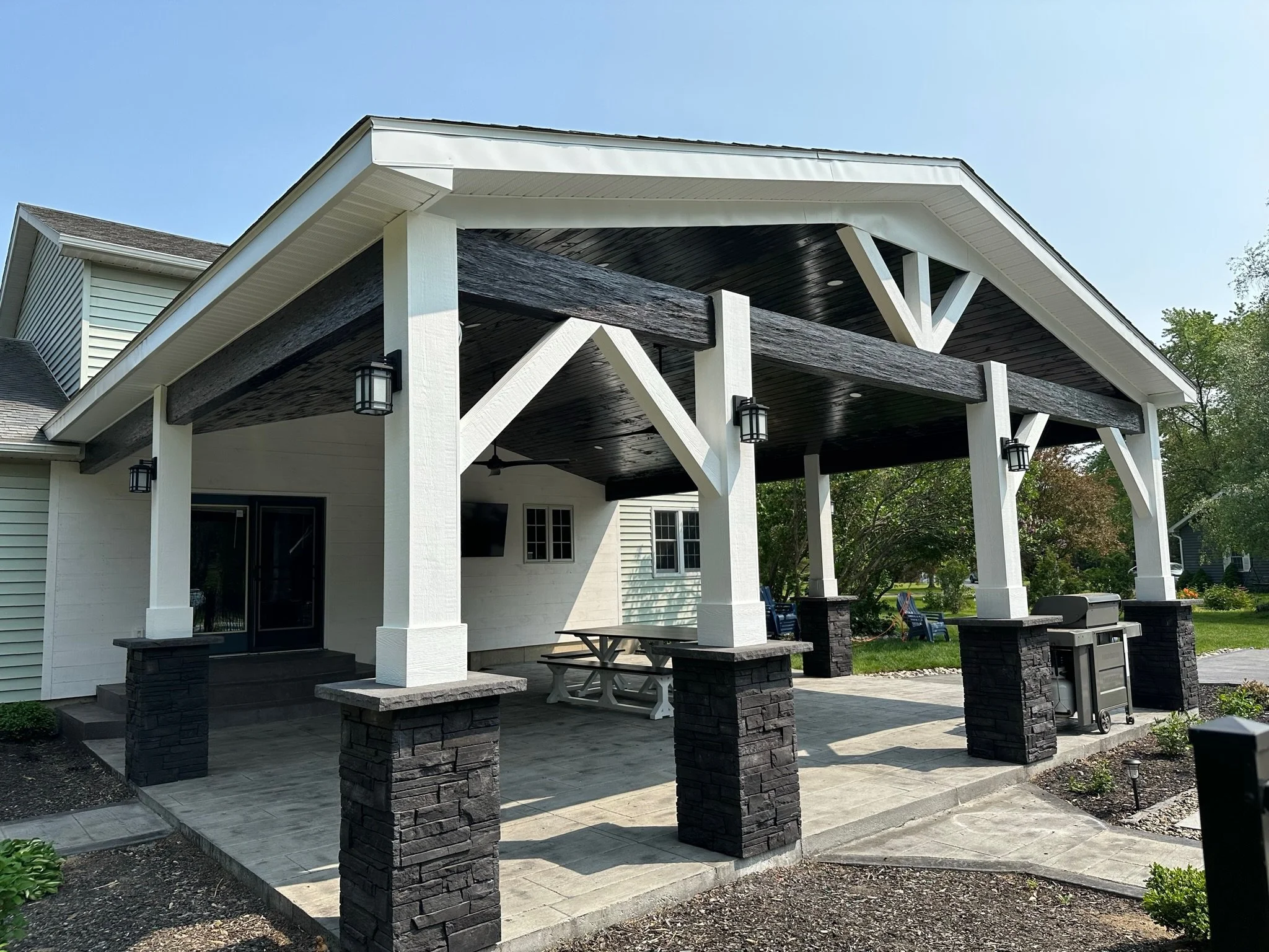 A backyard patio with a covered pergola featuring white and dark wood beams, black lantern-style lights, a seating area with a picnic table, and a built-in grill, with a lawn and trees in the background.