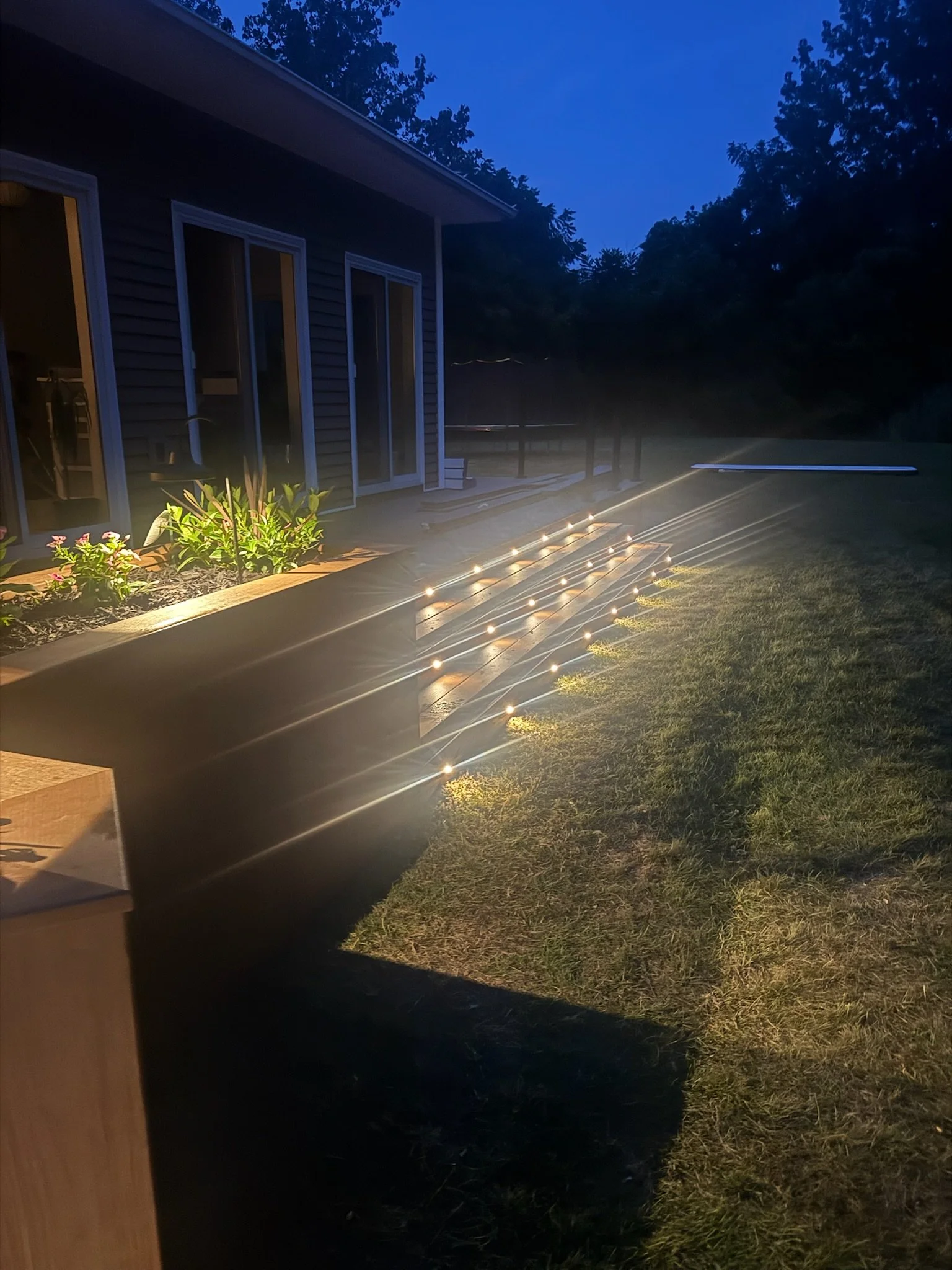 Outdoor scene at dusk showing a house with sliding glass doors, a garden bed with flowers, and a lawn with scattered solar-powered garden lights illuminating the area.