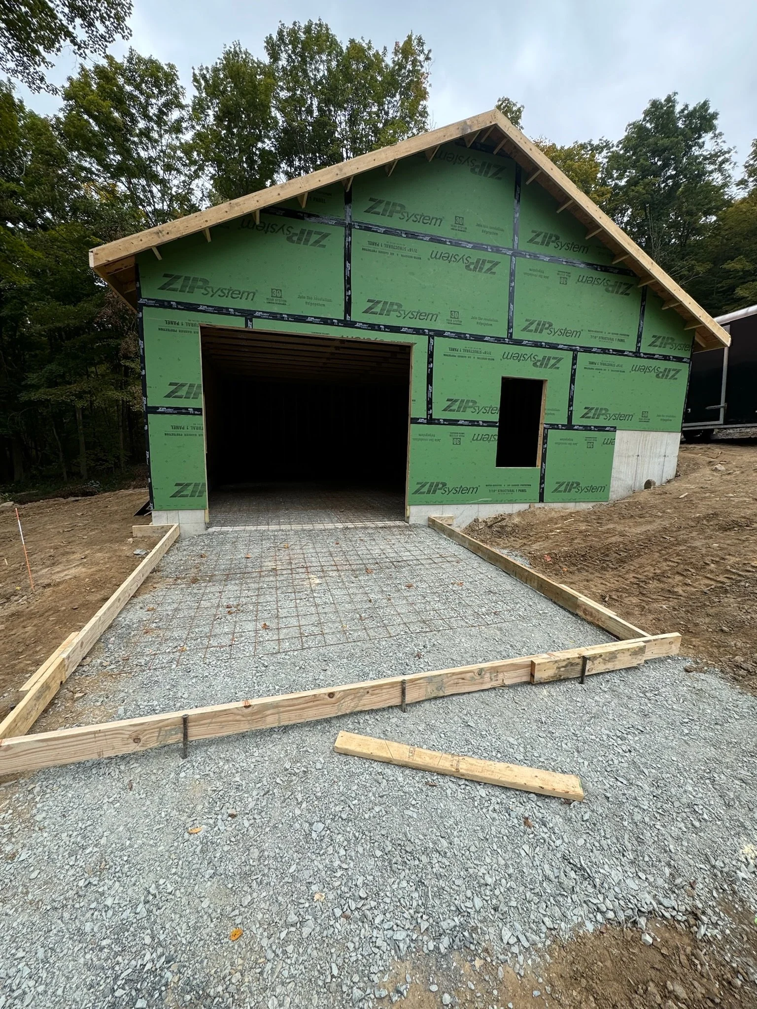 A house under construction with green building wrap, an incomplete driveway, and surrounded by trees.